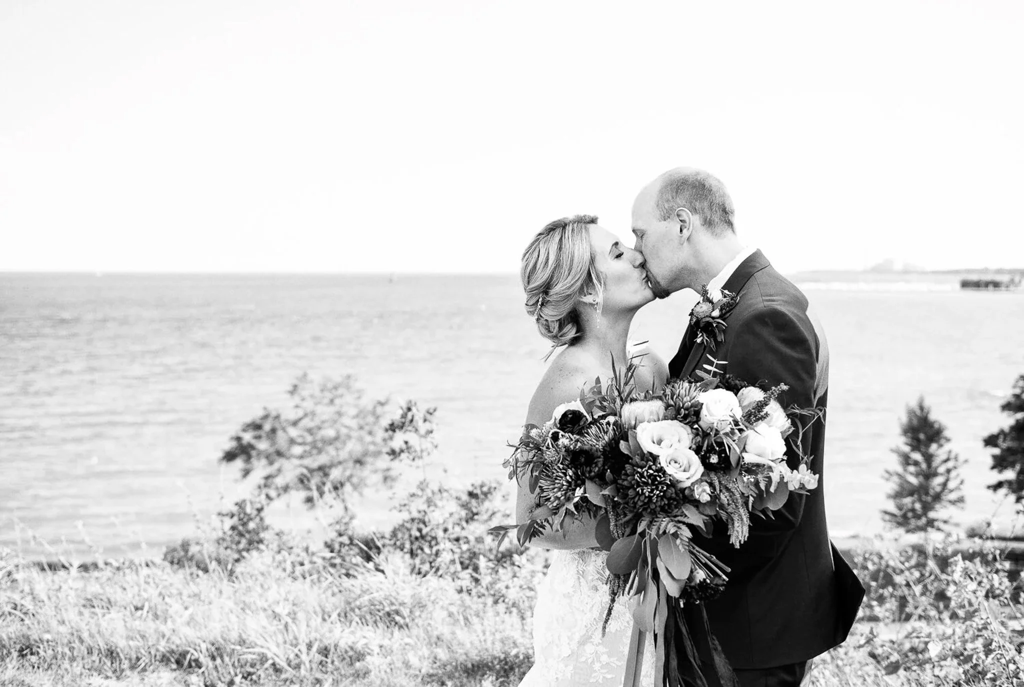 Black and white lakefront kiss with dramatic cascading bouquet of roses and protea, wide open Lake Michigan bay behind couple — Milwaukee wedding photography