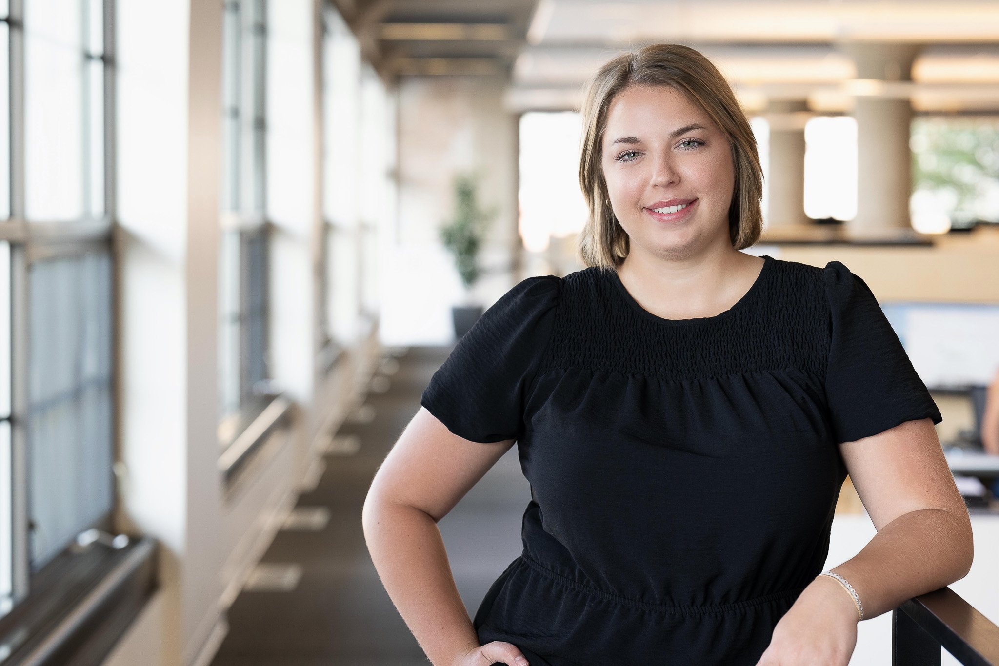 Professional woman headshot, black smocked short-sleeve blouse, smiling in open corporate office with large windows and interior columns — Milwaukee corporate headshot photography