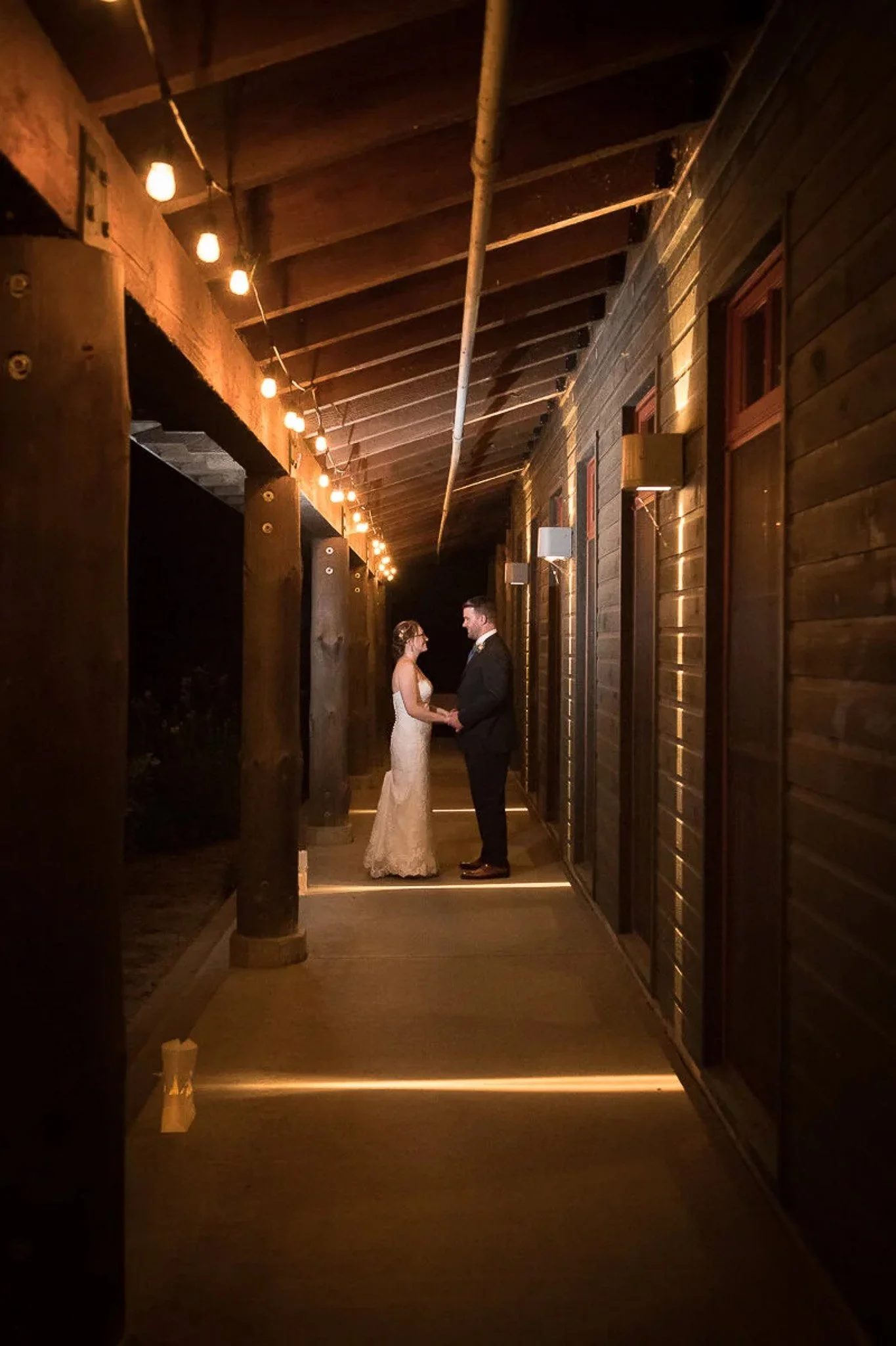 Dark moody barn corridor at night with string lights running along the ceiling, couple facing each other holding hands at the end of the hallway — Milwaukee wedding photography