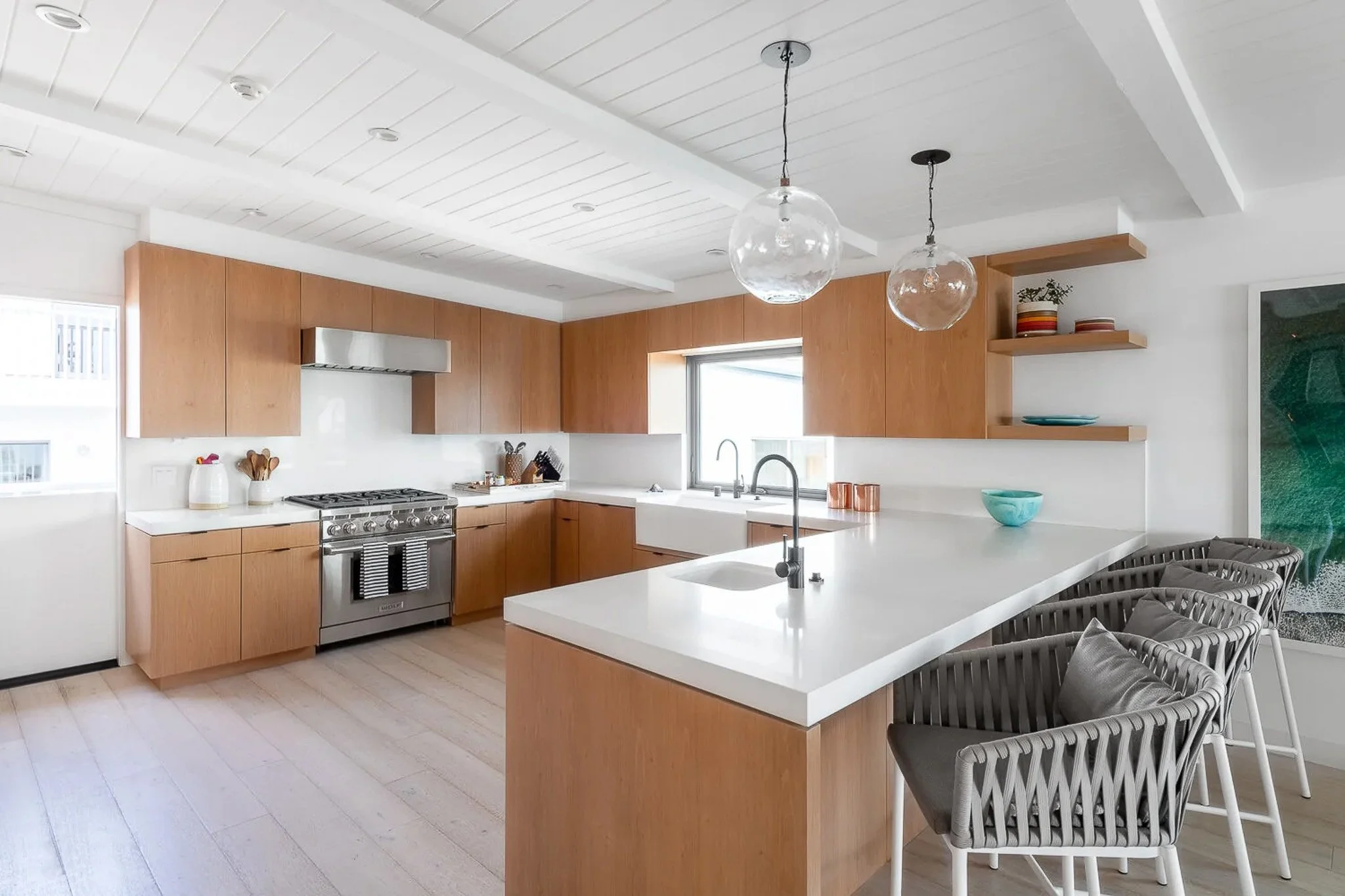 Mid-century modern kitchen with warm wood flat-panel cabinets, exposed painted ceiling beams, globe pendants, and wicker bar stools — California residential interior photography