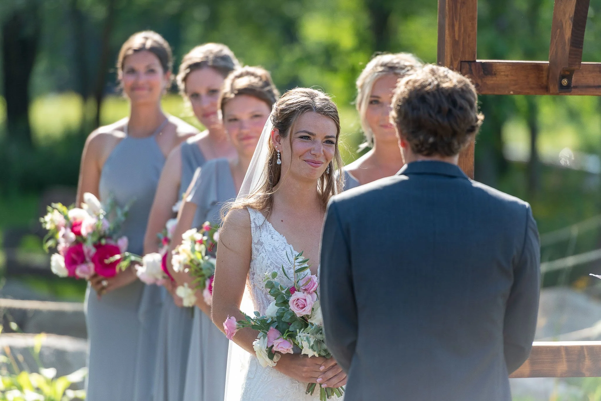 Outdoor ceremony moment, bride smiling at groom through wooden arch, dove-grey bridesmaids with pink and magenta bouquets visible in background, lush green trees — Milwaukee wedding photography