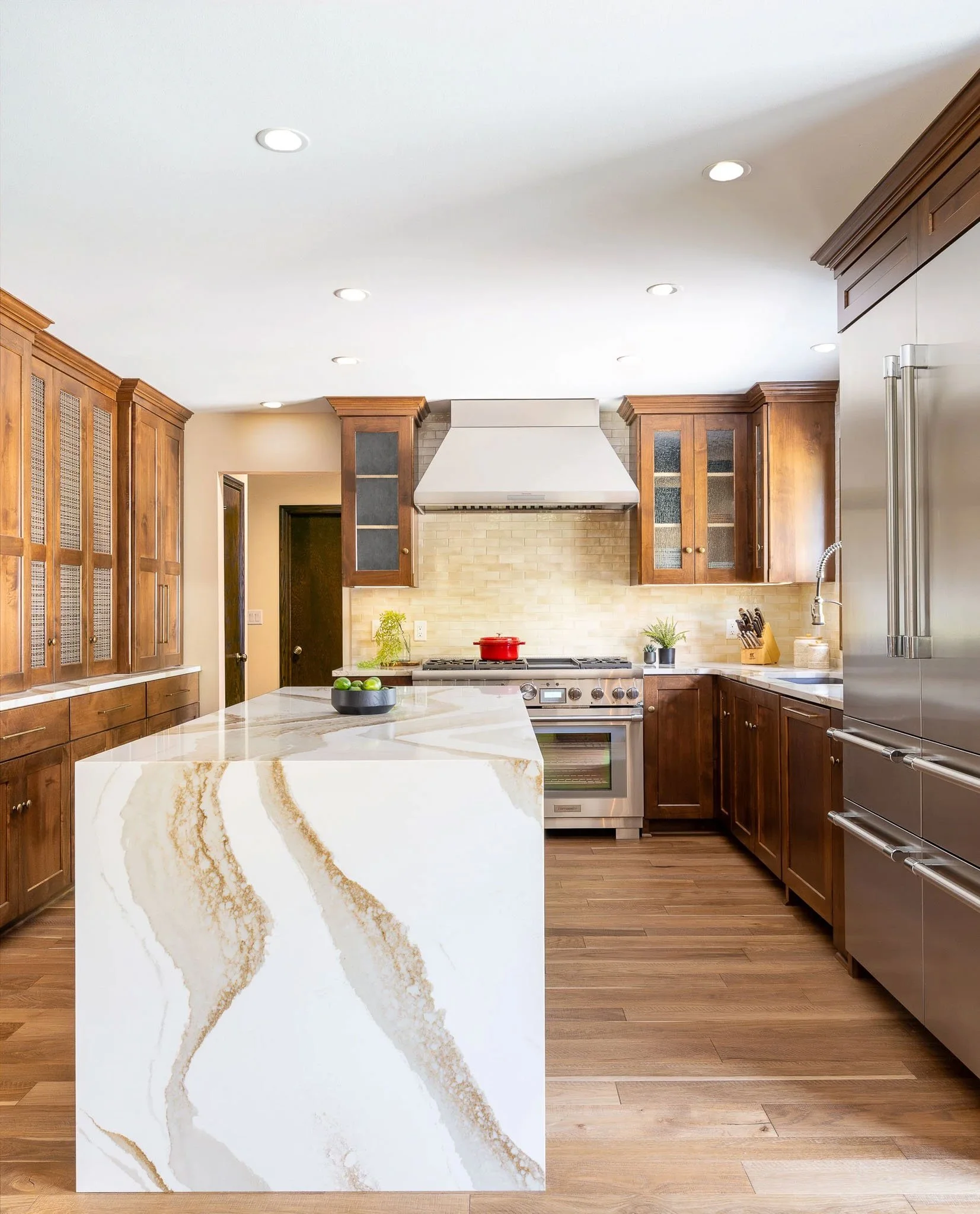 Dark walnut kitchen with dramatic gold-veined white marble waterfall island, glass-front upper cabinets, stainless professional range with red Dutch oven, and Sub-Zero refrigerator — Milwaukee Brookfield residential interior photography