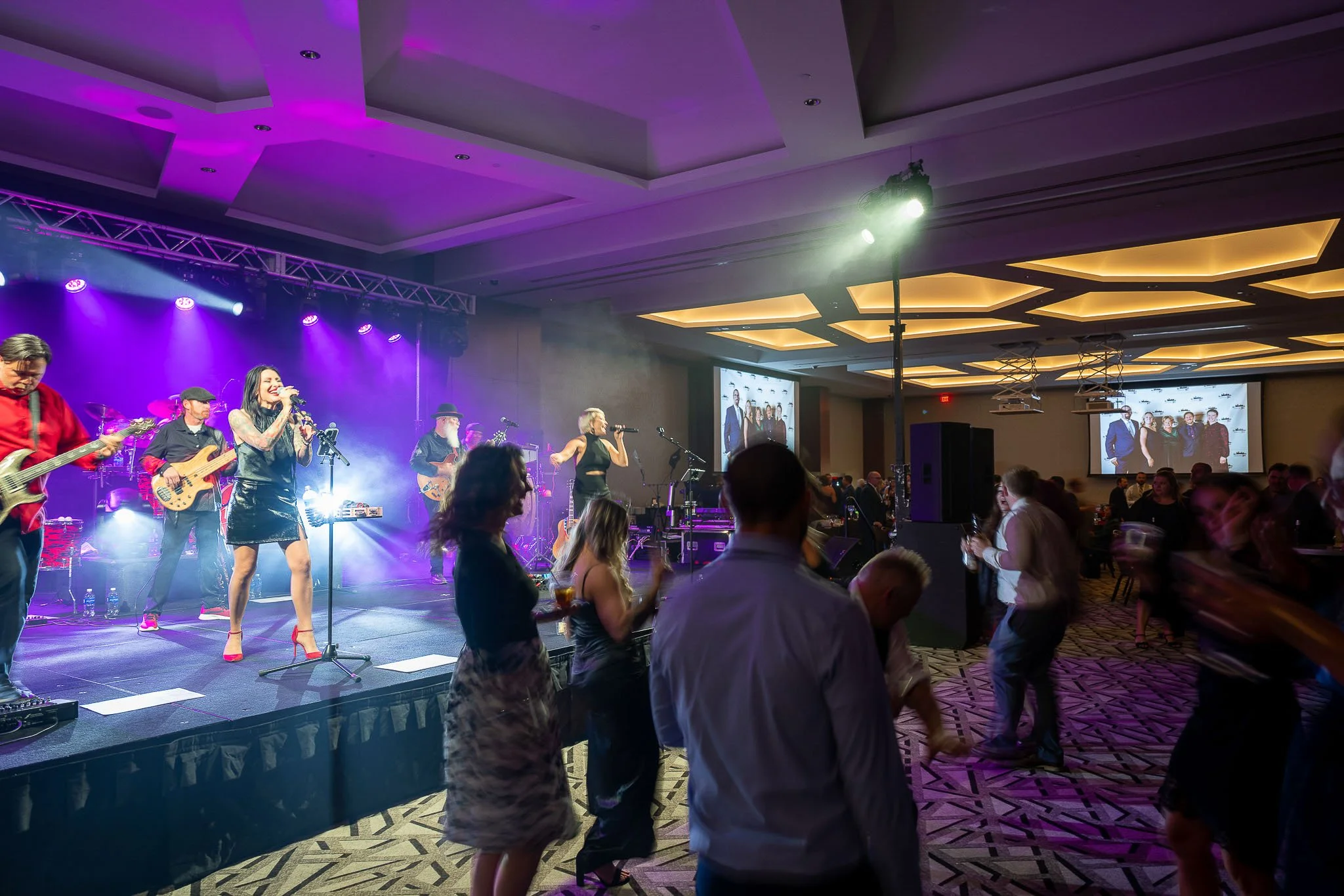 Live band performing at hotel ballroom gala, female singer in black outfit and red heels at microphone, guests dancing under purple stage lighting with projection screens — Milwaukee Sprinkler Fitters Local 183 event photography