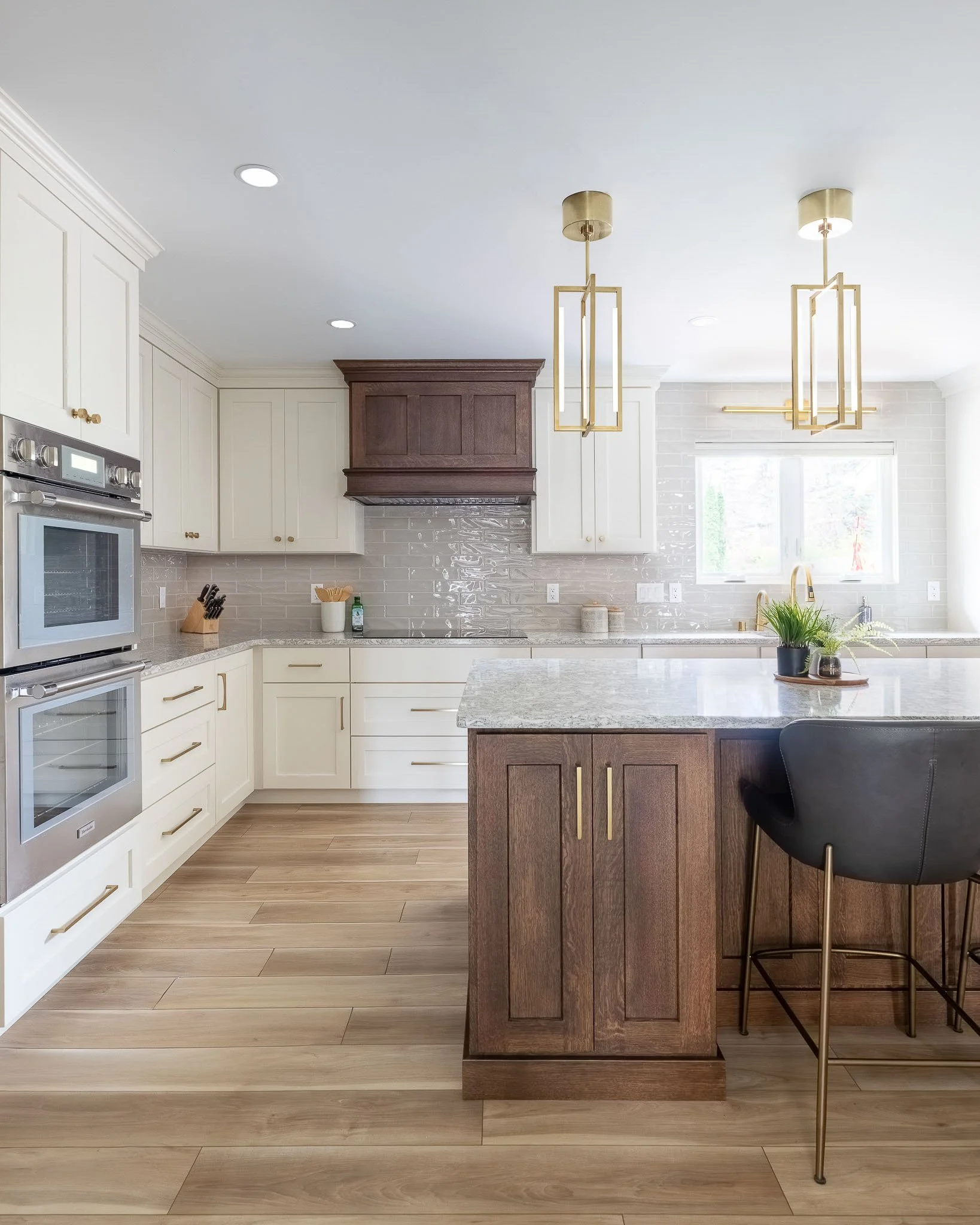 Two-tone kitchen with cream upper cabinets, dark walnut island and range hood, iridescent wavy tile backsplash, grey-green granite countertops, and gold lantern pendants — Milwaukee Brookfield residential interior photography