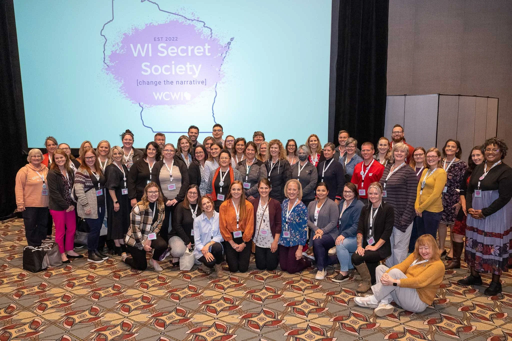 Large group photo at WI Secret Society conference in front of "WI Secret Society" projection screen, attendees with lanyards and name badges — Milwaukee Wisconsin conference event photography