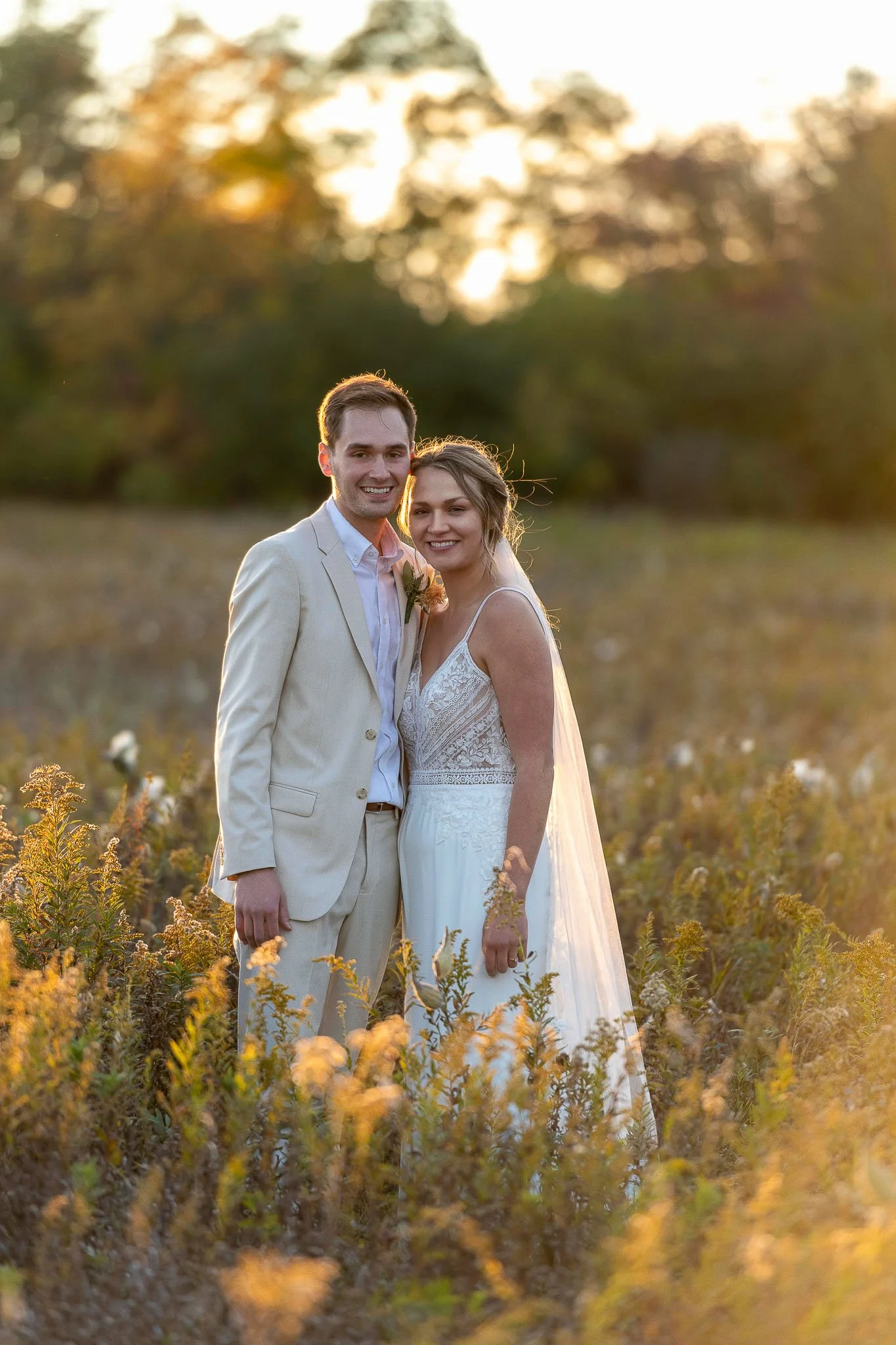 Couple standing in golden wildflower field at sunset, groom in cream linen suit, bride in boho lace spaghetti-strap gown, warm light through the trees — Milwaukee wedding photography