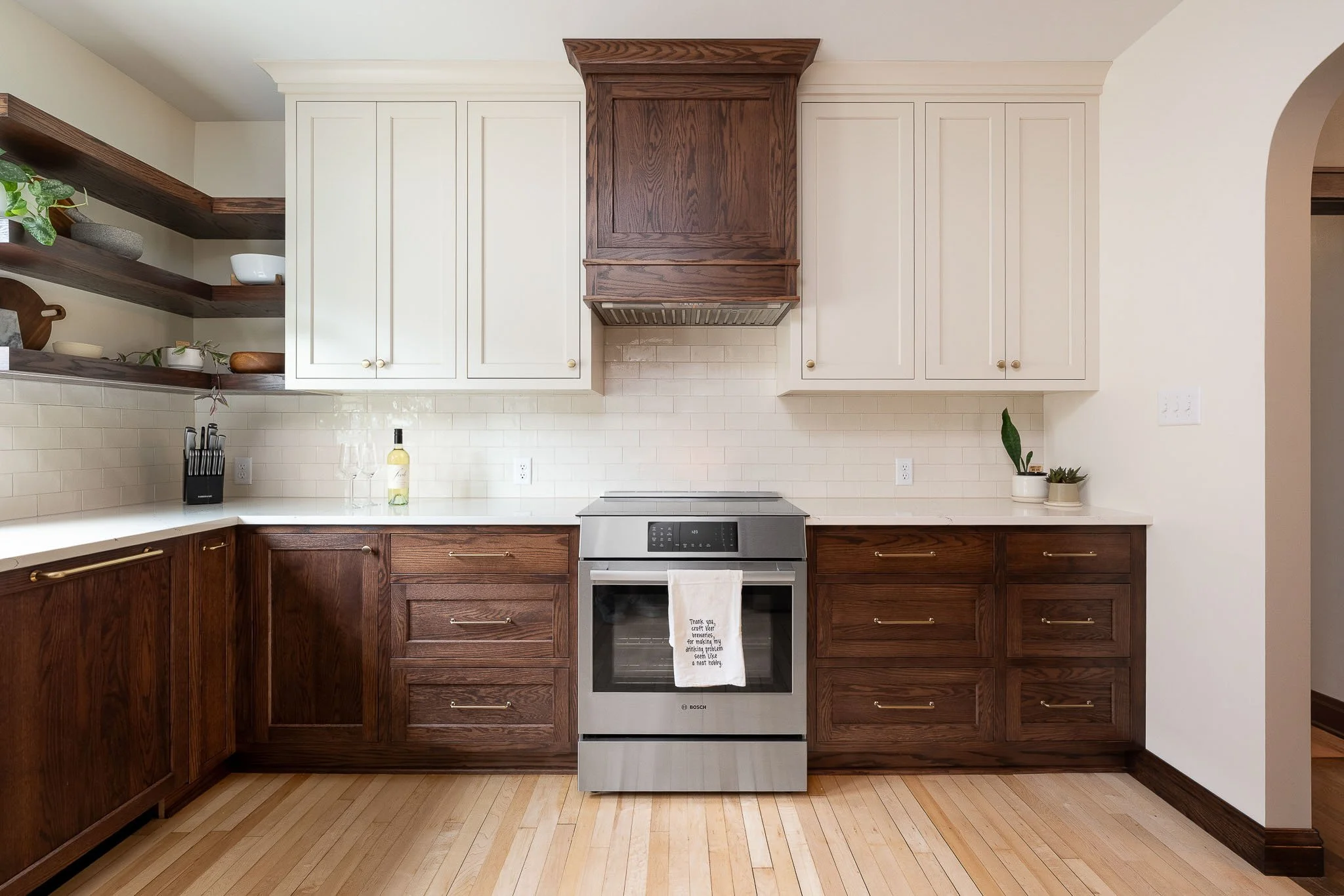Two-tone kitchen with cream upper cabinets, dark walnut lower cabinets, walnut range hood, cream subway tile backsplash, and floating wood shelves with plants — Milwaukee residential interior photography