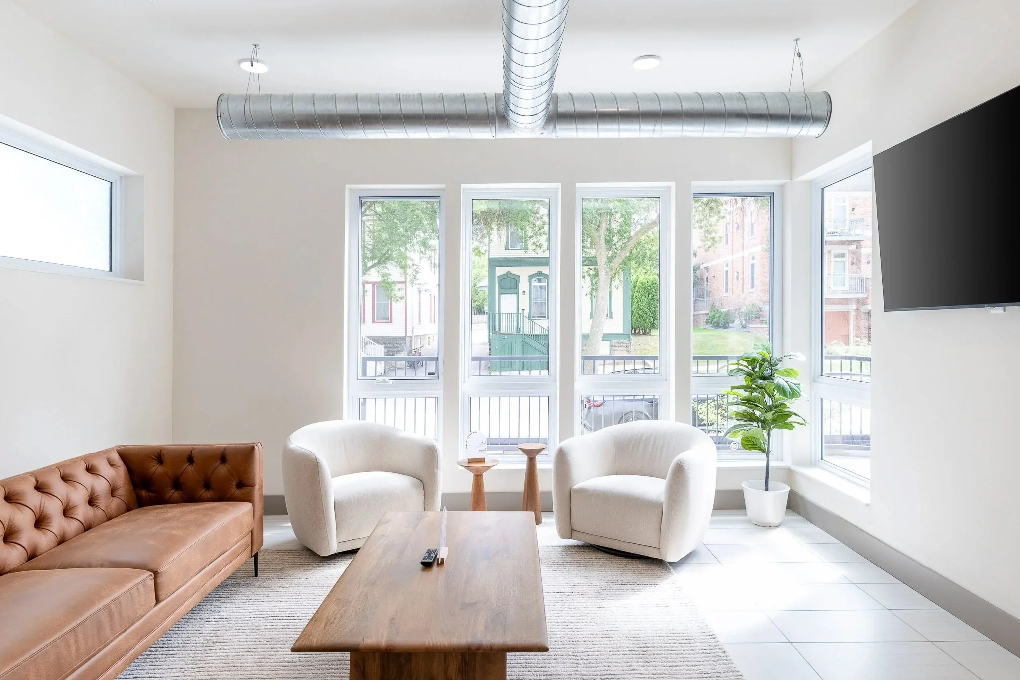 Modern open living room with exposed ductwork, four tall narrow windows overlooking brick street, tufted leather sofa, and white barrel chairs — Milwaukee residential interior photography