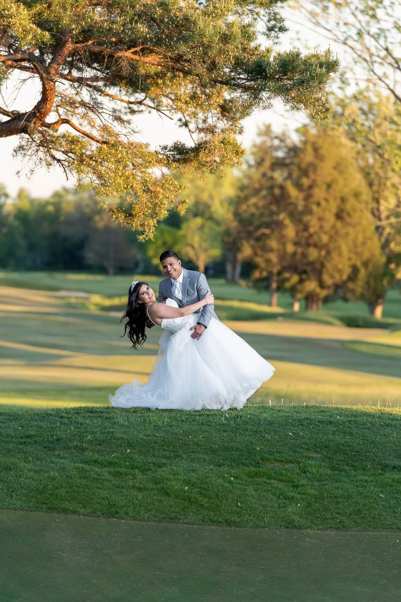 Golf course couple dip at golden hour, bride in full white ball gown with tiara, groom in grey suit, golden pine tree overhead — Milwaukee wedding photography