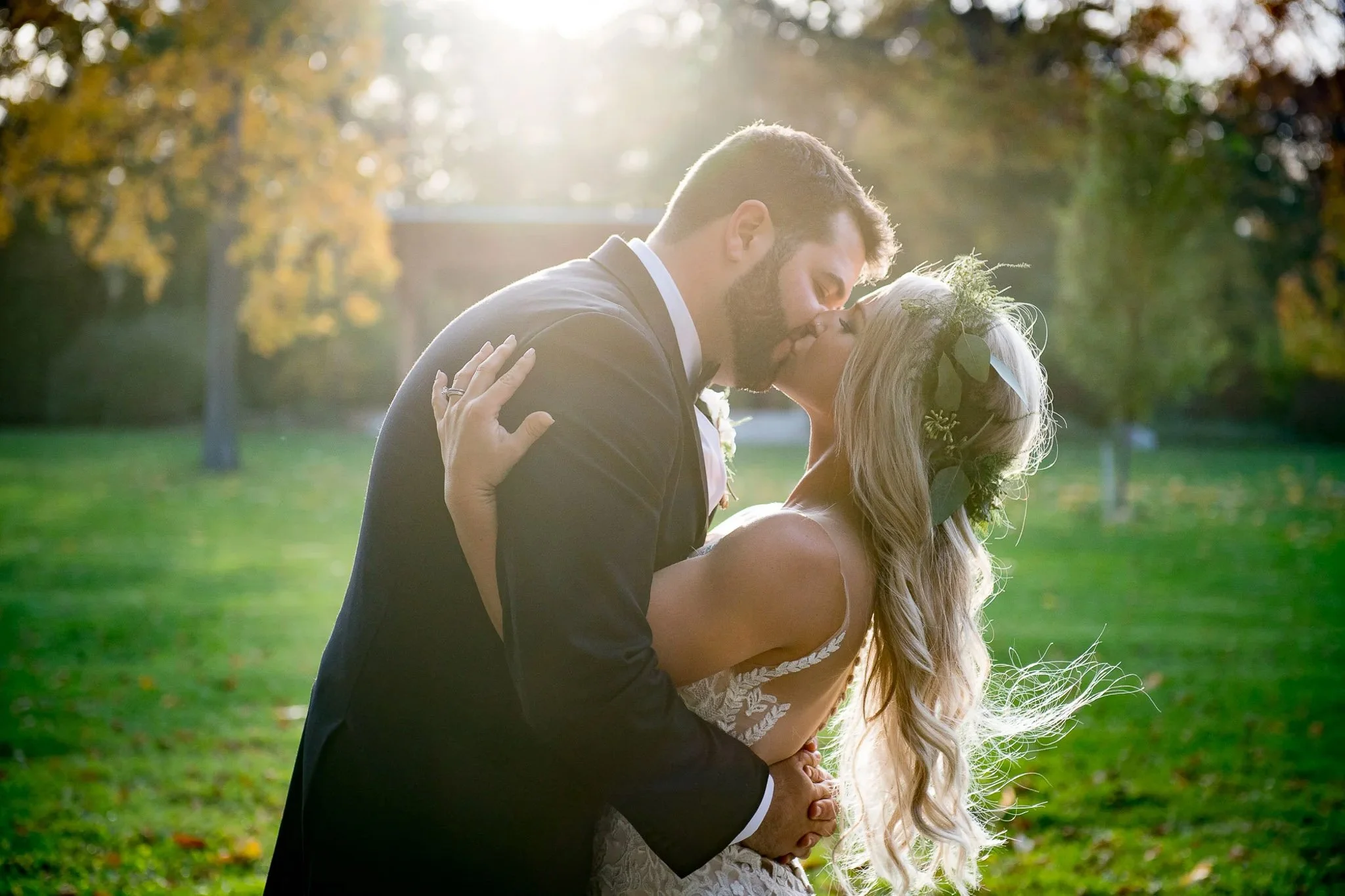 Golden autumn backlit kiss in park, bride with greenery floral crown and flowing waves, groom in black tuxedo, yellow autumn trees surrounding — Milwaukee wedding photography