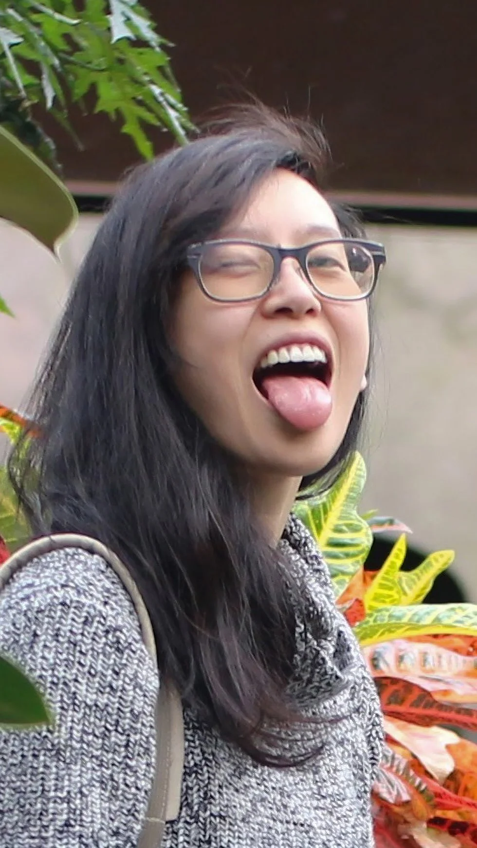 Rachel, a Vancouver and Richmond-based improv comedian and part of the improv comedy troupe 'Those Types' with long dark hair, glasses, and a patterned sweater making a funny face with her mouth open and tongue out, outdoors near green plants.