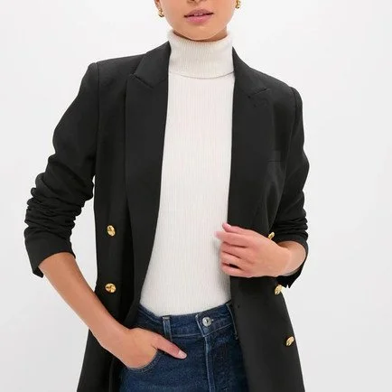 Woman wearing a black blazer with gold buttons over a white turtleneck, with one hand in her pocket and the other holding her blazer.