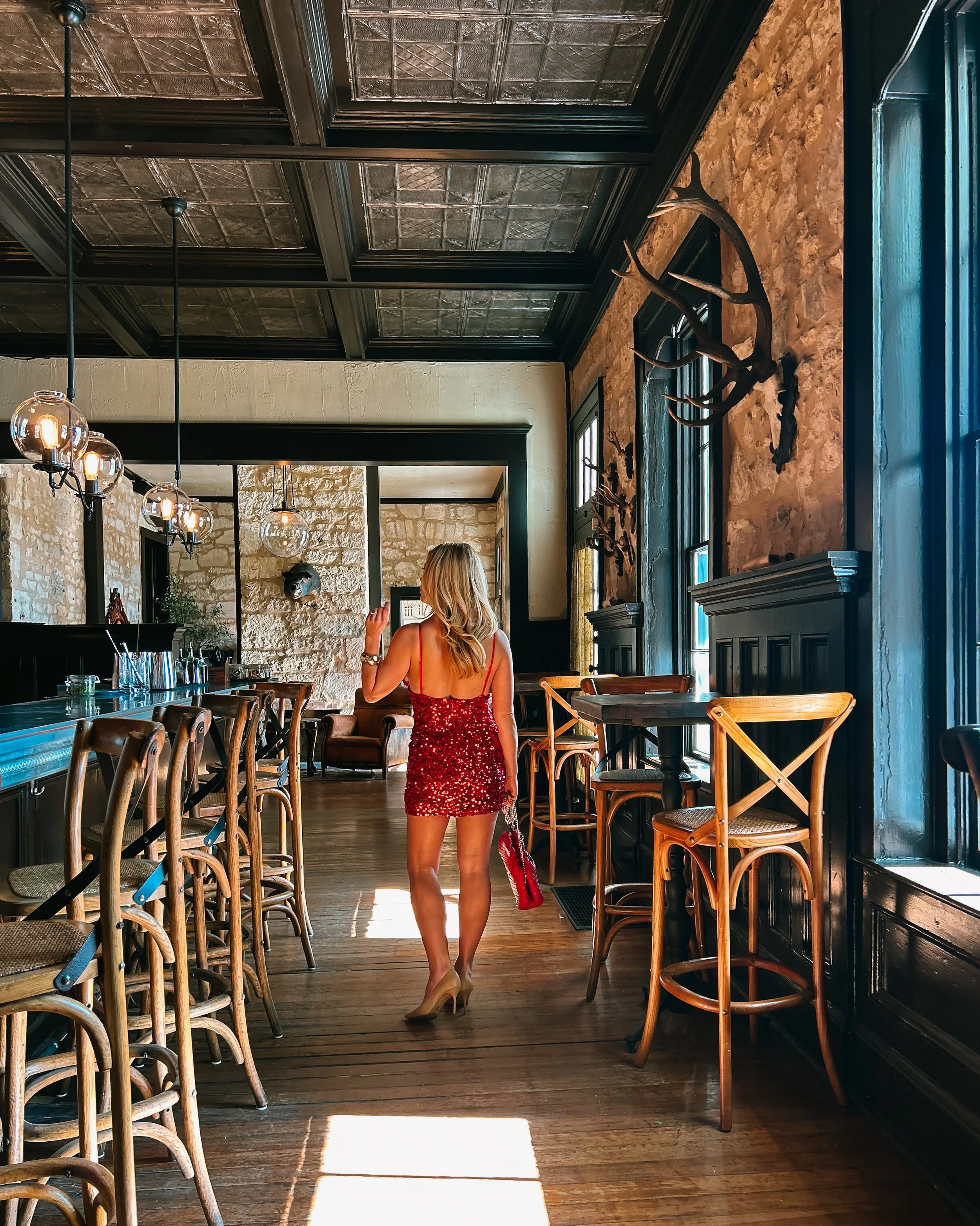 A woman with long blond hair wearing a red sequined dress and nude high heels walking through a rustic restaurant interior featuring wooden chairs, tables, stone walls, deer antlers mounted on the wall, and large windows with dark trim.
