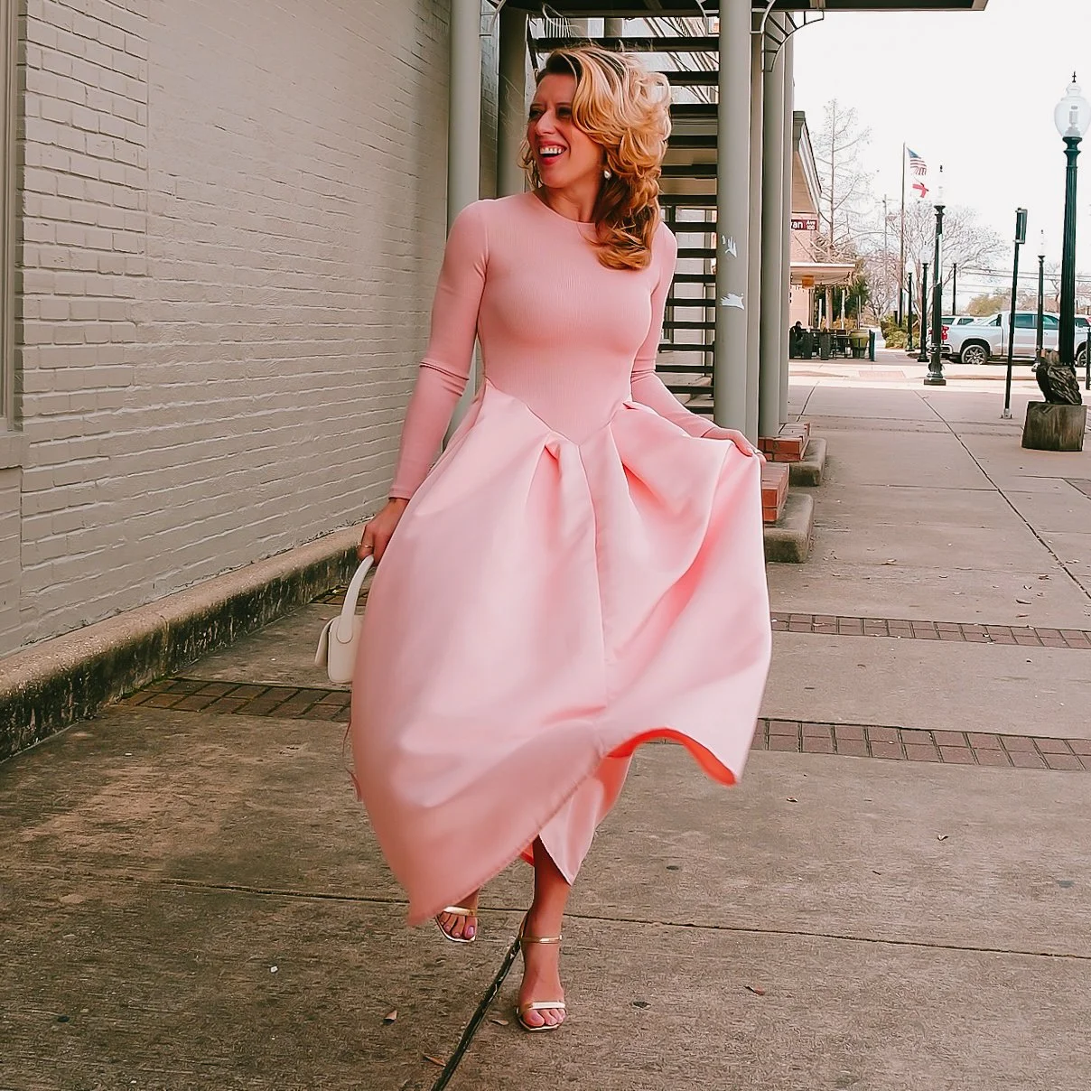 Woman in a light pink dress walking outdoors on a sidewalk, smiling and holding a small matching purse.
