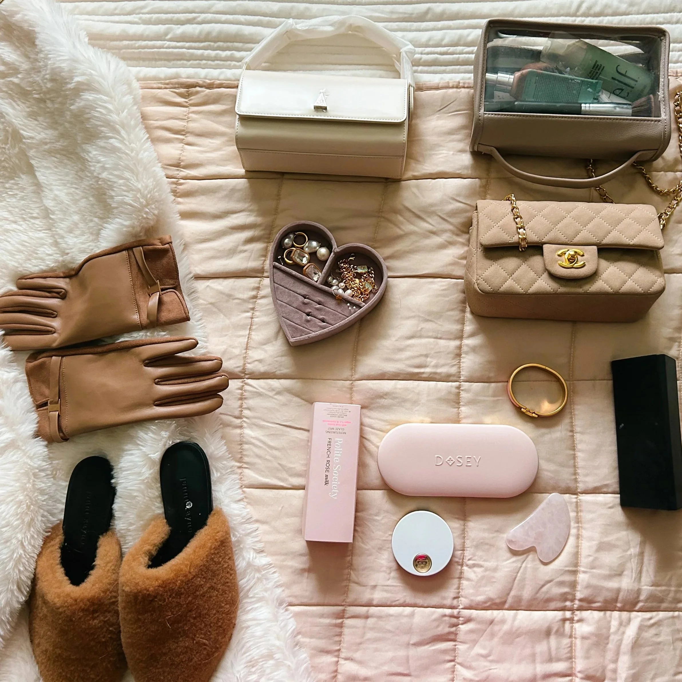 A collection of beige and brown accessories including handbags, a fluffy white blanket, makeup brushes, jewelry, and a pair of slippers arranged on a bed with a quilted beige cover.