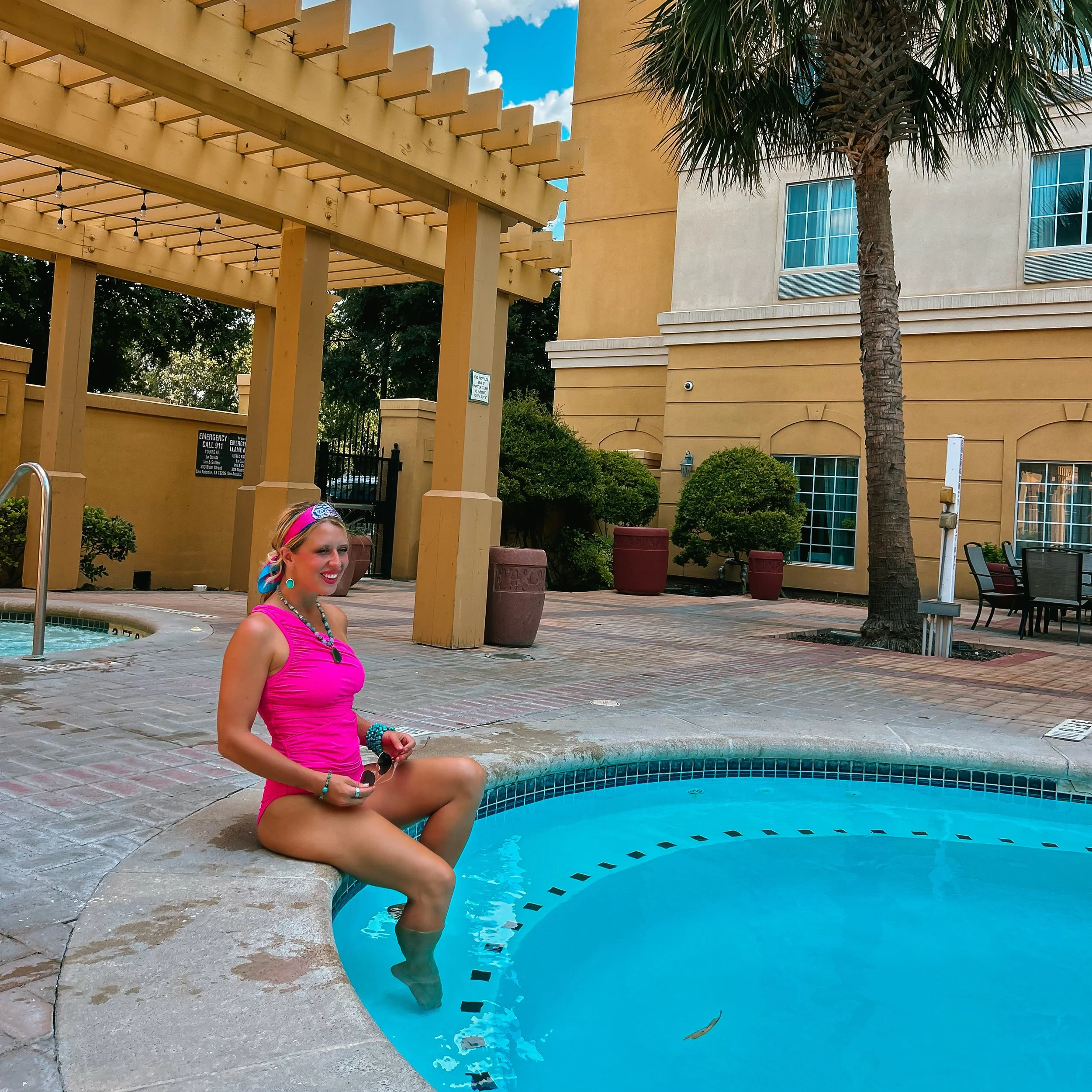A woman in a bright pink top and colorful headband sitting poolside with her feet in the water, smiling, with a yellow building, palm tree, and blue sky in the background.