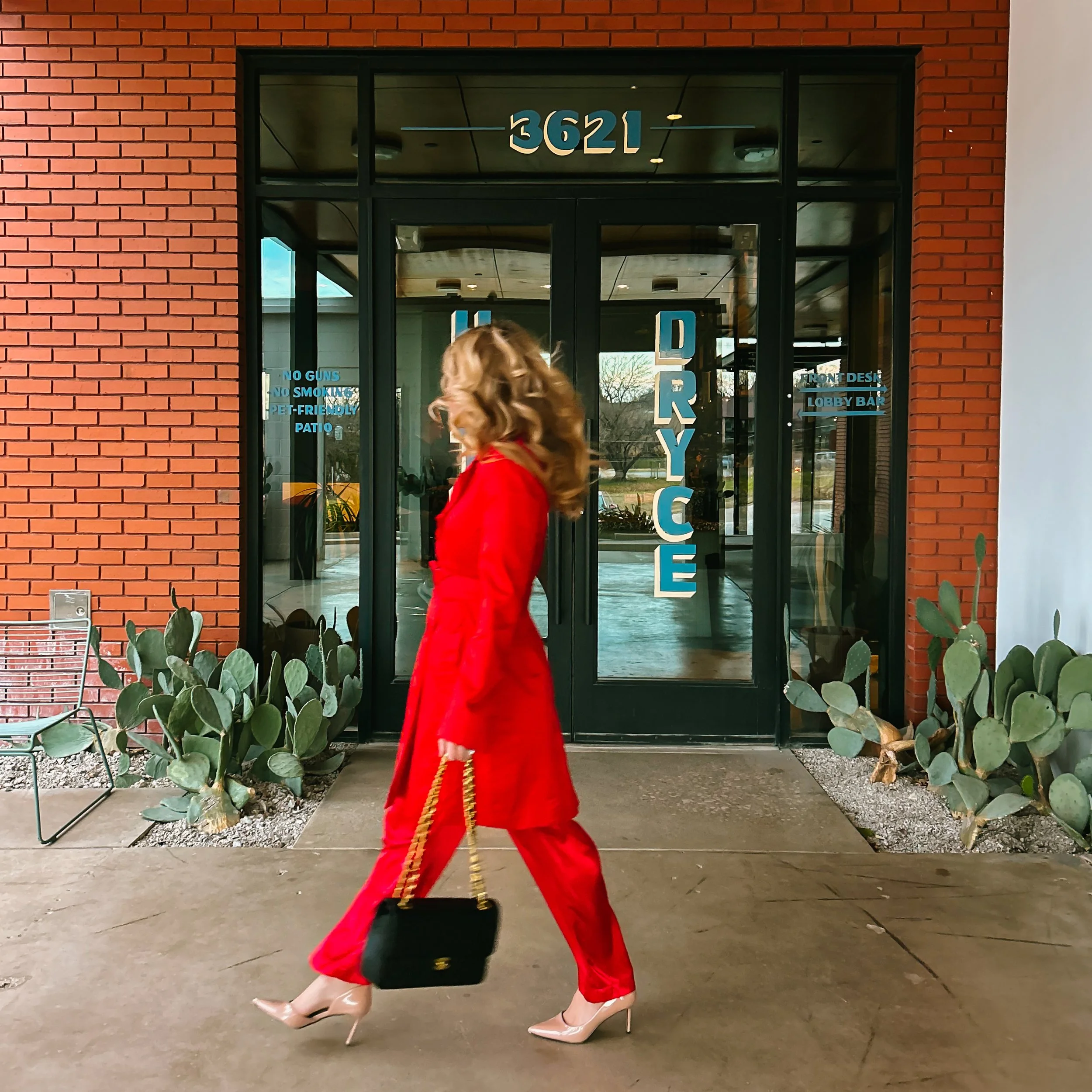A woman with long curly hair dressed in a red coat and red pants is walking inside a building with a red brick exterior. She is wearing beige high heels and carrying a small black purse with a gold chain. The building has a glass door with blue lettering, and the number 3621 is displayed above the door. There are plants with large green leaves near the entrance, and a metal bench and a small electrical box on the brick wall nearby.