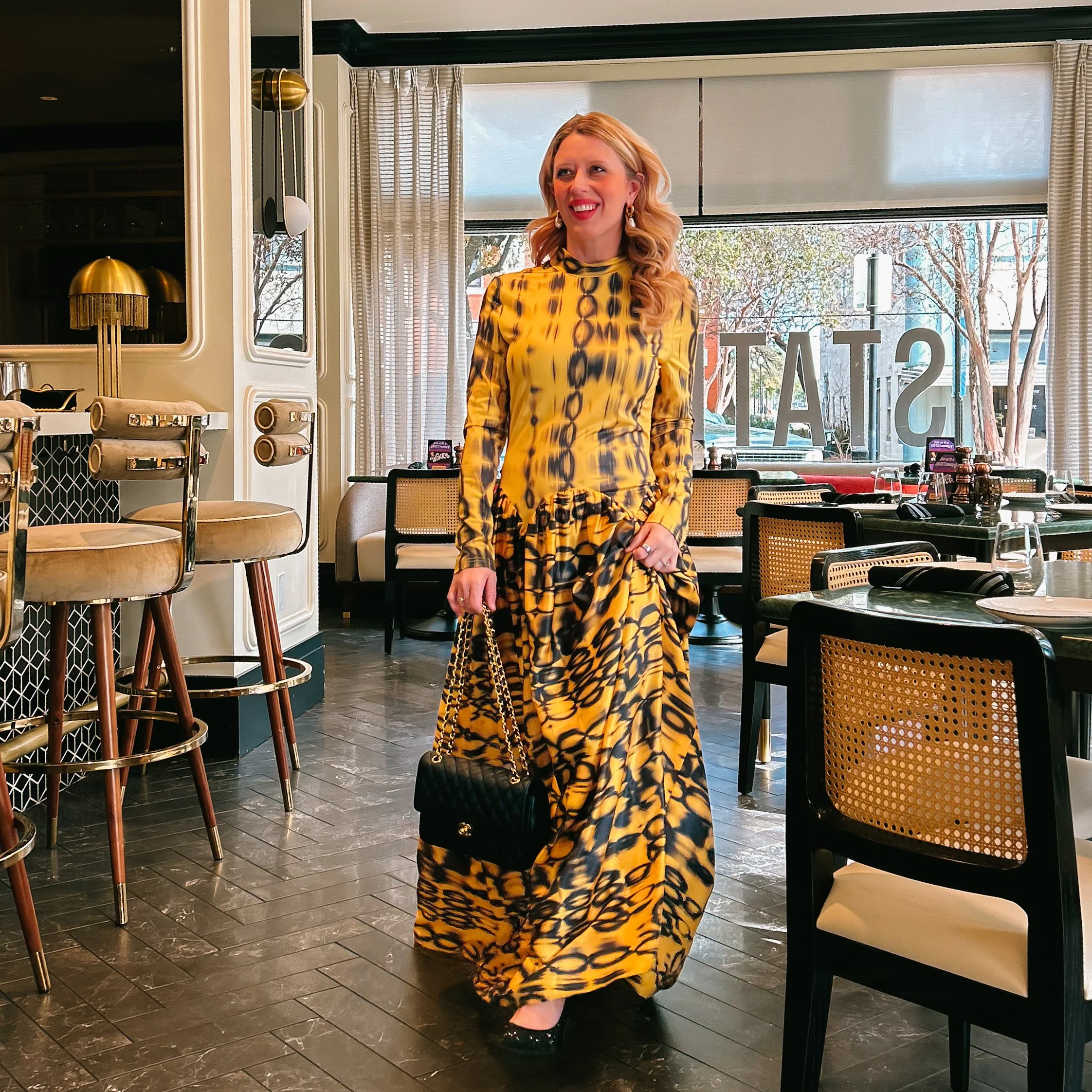 A woman standing inside a restaurant, wearing a yellow and black patterned dress with ruffled hem, holding a black quilted handbag with gold chain strap, smiling, with a dining area and large window behind her showing trees outside.