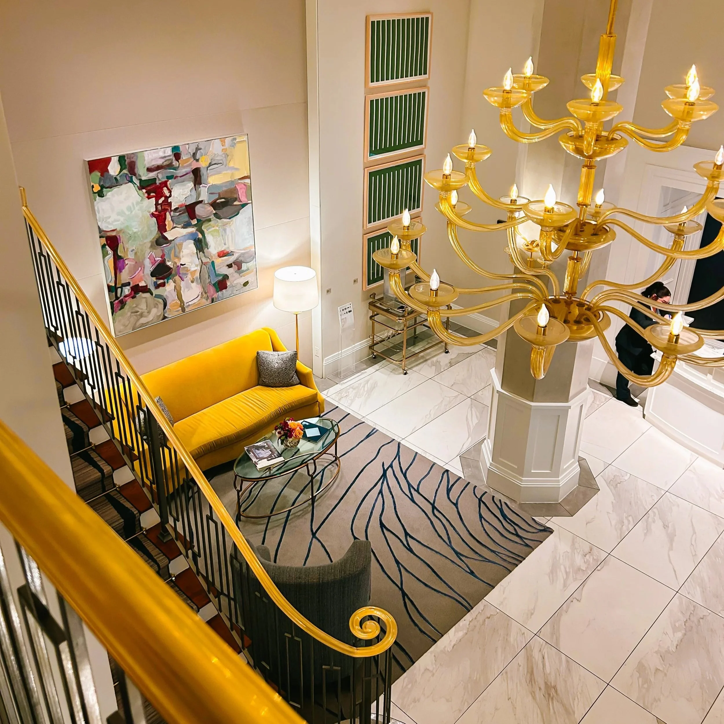 Interior view of a hotel lobby showing a large golden chandelier, abstract artwork on the wall, a white table lamp, a green and gold hallway runner, a yellow sofa with a gray pillow, and a black and gold side table with a flower arrangement.