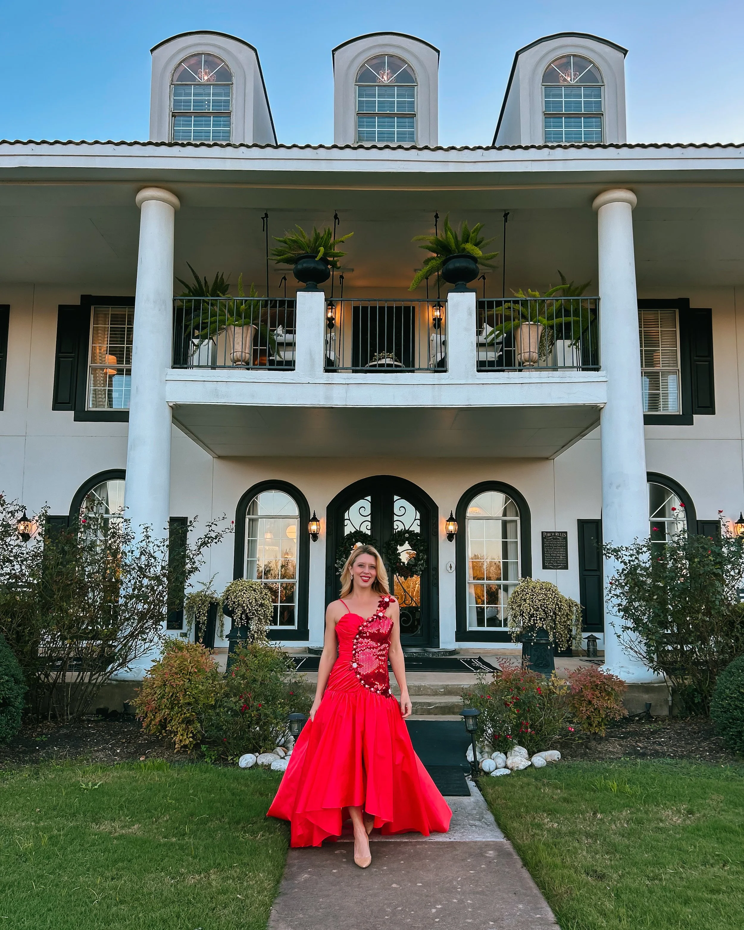 A smiling woman in a red dress stands on a concrete pathway in front of a house with a large porch and columns. The house has three attics with arched windows and a decorated front door.