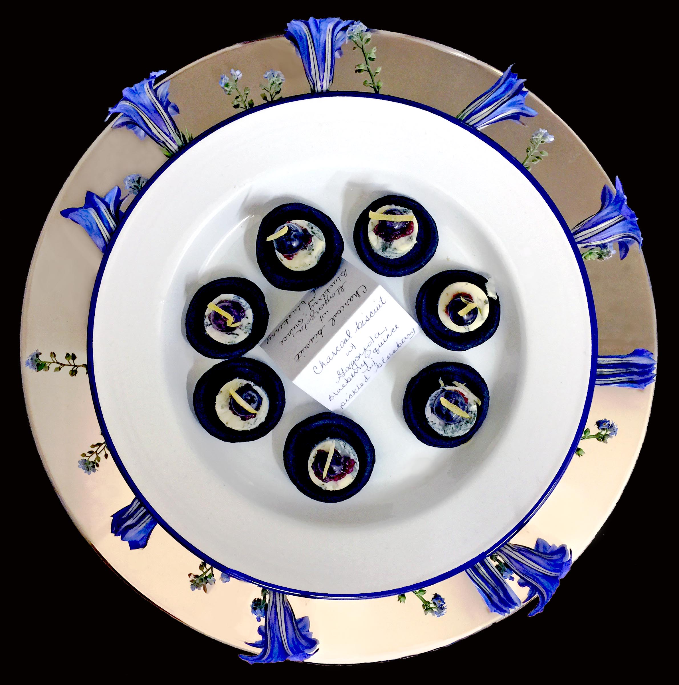 Tray of charcoal biscuit canapés served on an enamel plate within a sterling silver inset plate and decorated with fresh forget-me-not and gentiana flowers.