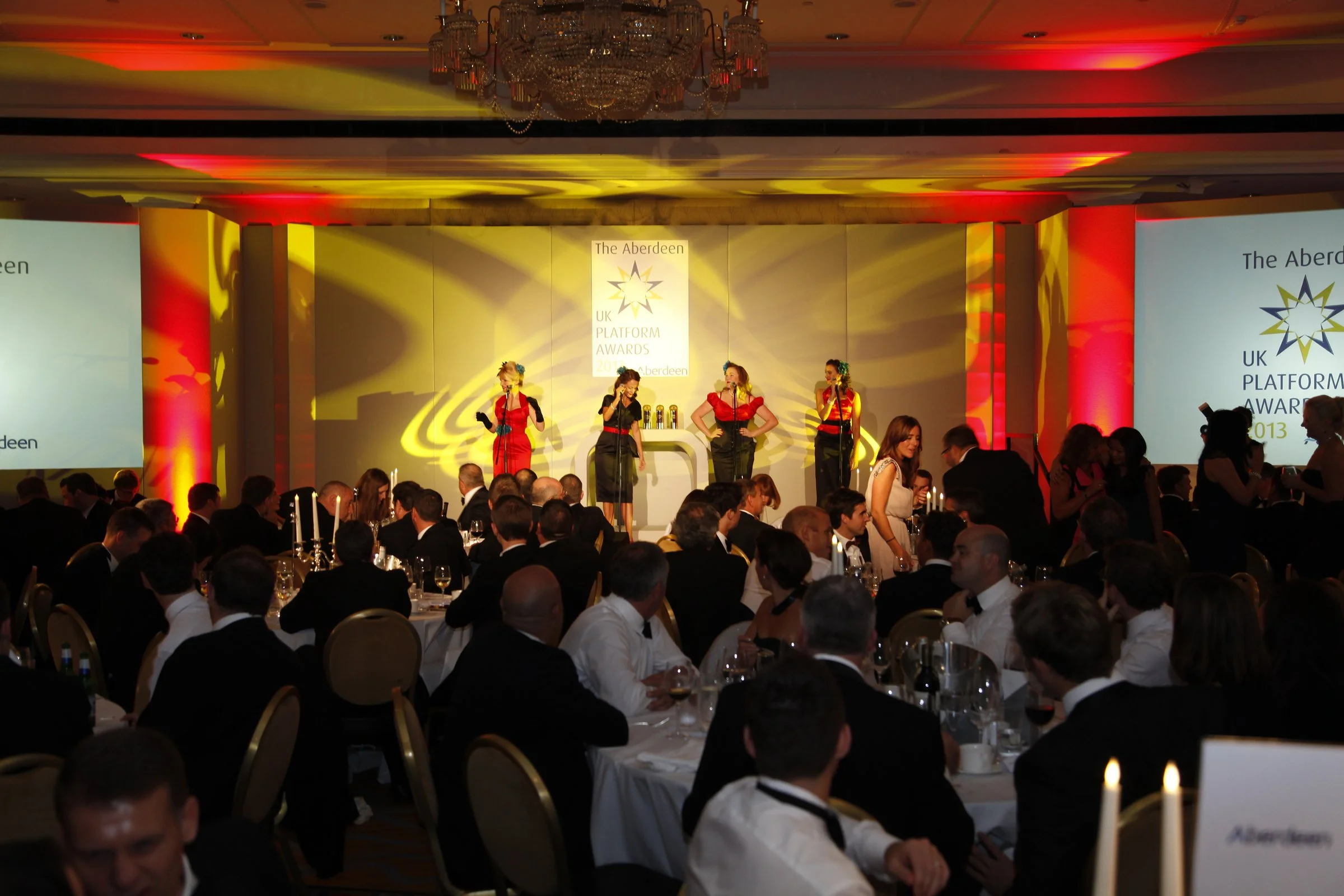 4 Ruby Dolls perform on stage at a corporate event in a room set out with people having dinner and drinks