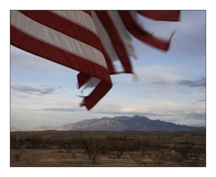 A US flag tethered to a family gatepost flutters in the afternoon breeze on the road between Arivaca and Amado, in the background is the Santa Clara mountain range.