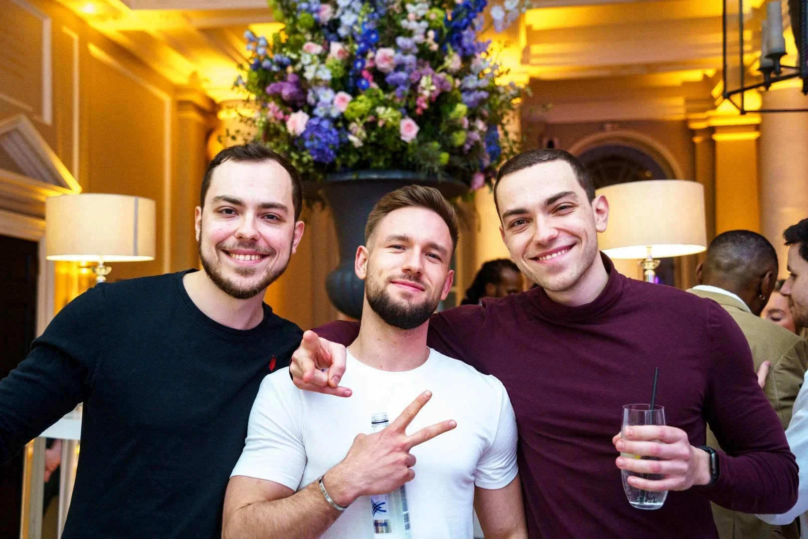 Three smiling young men standing close together indoors at a social gathering, with a large floral arrangement hanging above them. Two of them are making peace signs and holding drinks.