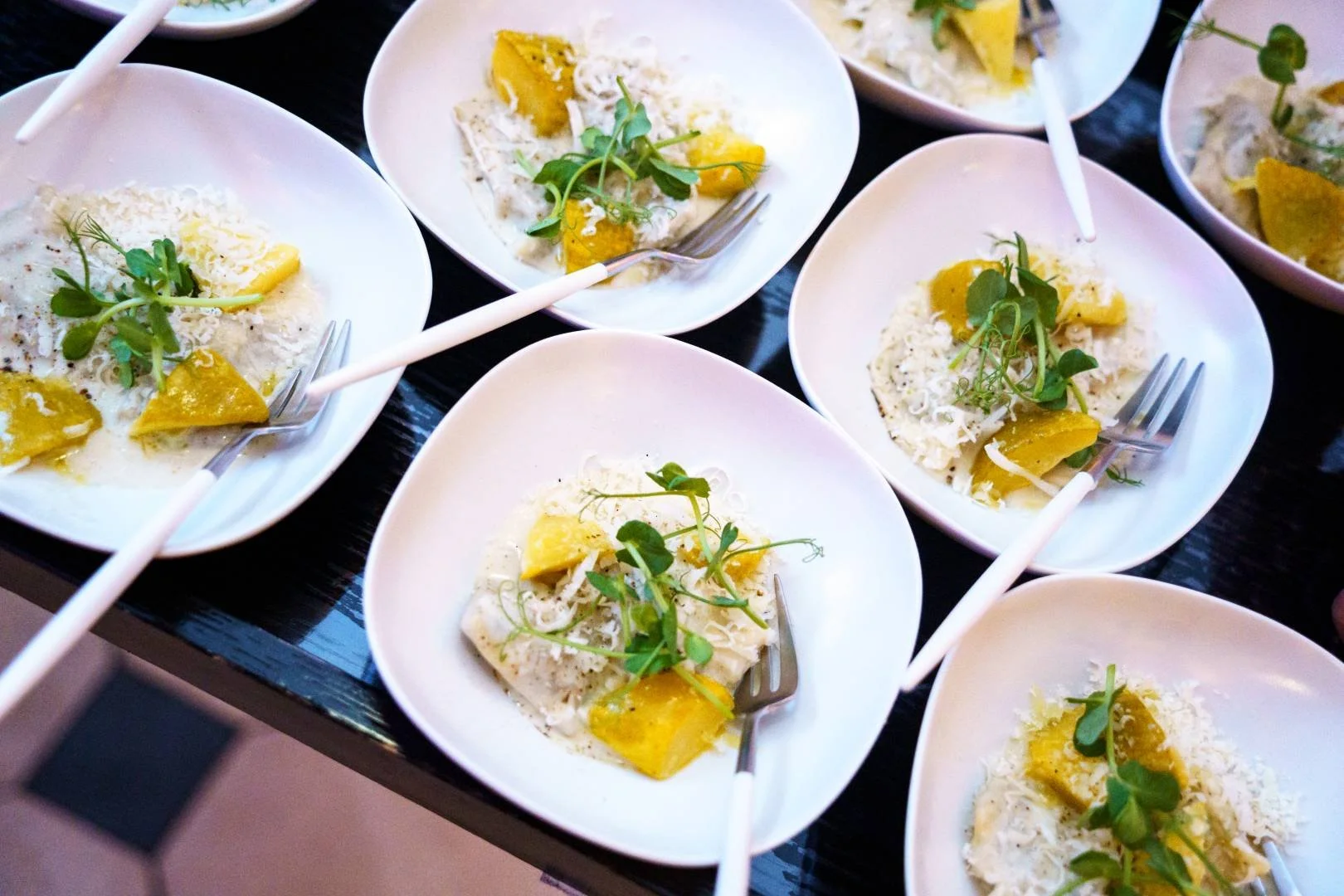 Multiple small white bowls containing a creamy dish garnished with shredded cheese, green herbs, and yellow sauce, with small forks inside some bowls on a dark surface.