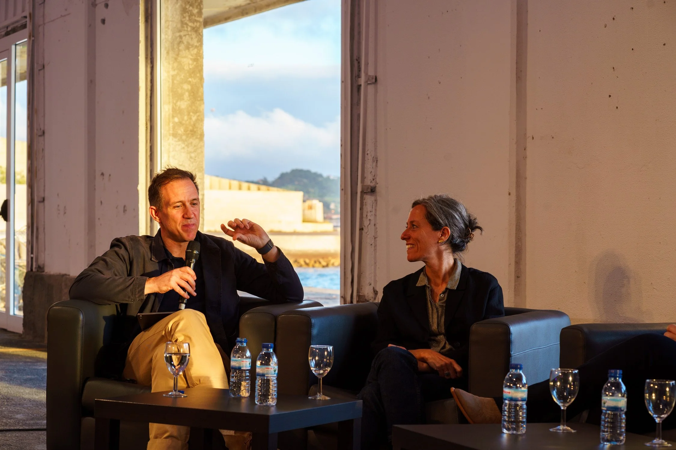 Two people are seated on black armchairs in a room with large open windows showing a water and hill view in the background. One man with short dark hair, wearing a dark shirt and beige pants, holds a microphone and gestures while speaking. A woman wi