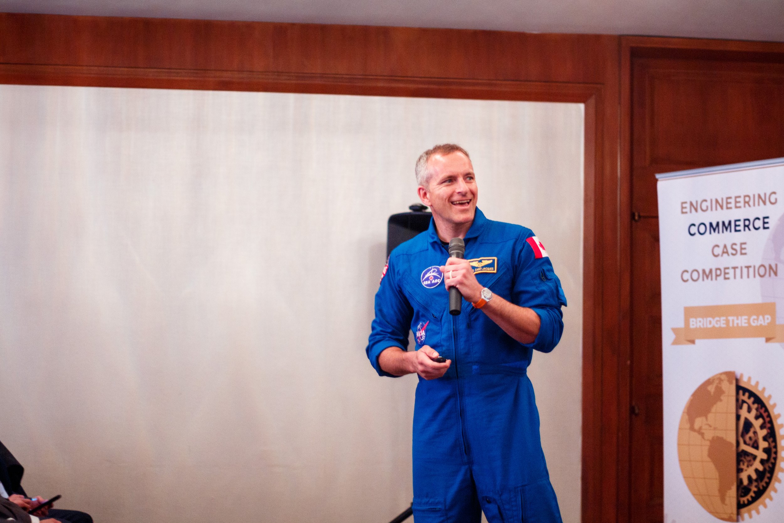 A man in a blue astronaut suit holding a microphone and smiling during a presentation.