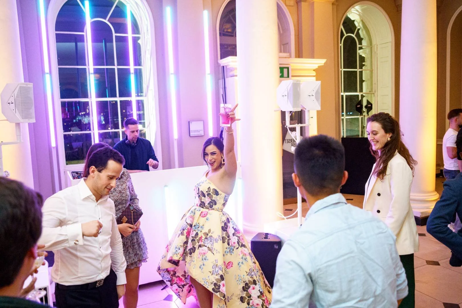 People dancing and socializing at an indoor party with colorful lighting, a woman in a floral dress holding a drink, others smiling and engaged in conversation.