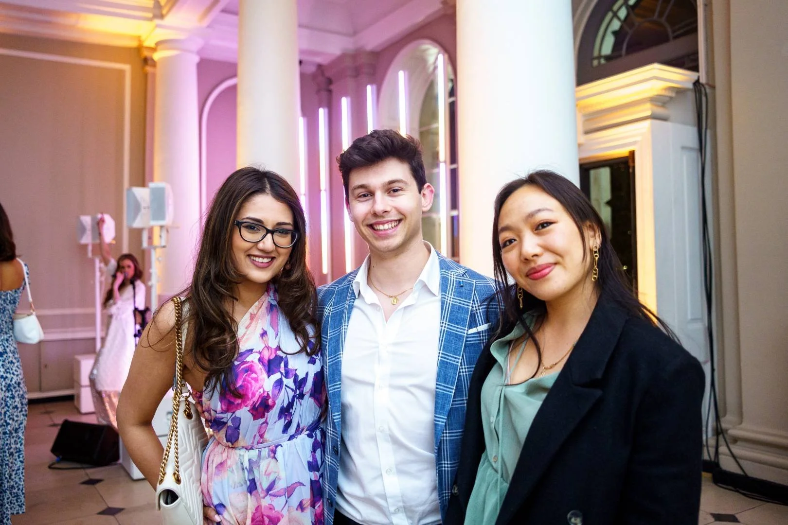 Three young adults smiling and posing together at an indoor event with elegant decor and pink lighting.