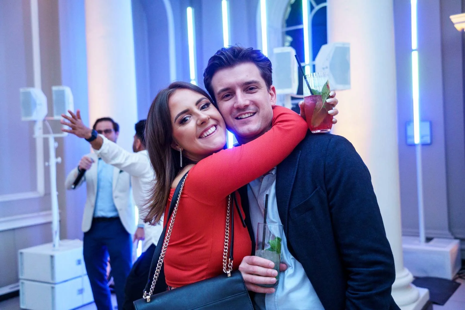 Young couple hugging and smiling at a party, holding cocktails, with a man in the background singing or speaking into a microphone.