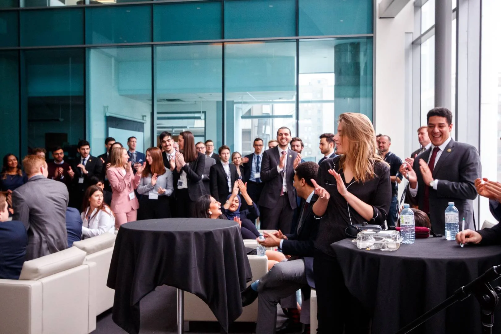 A group of business professionals at an indoor conference or networking event, some standing and some sitting, clapping and smiling, with large windows and a cityscape background.
