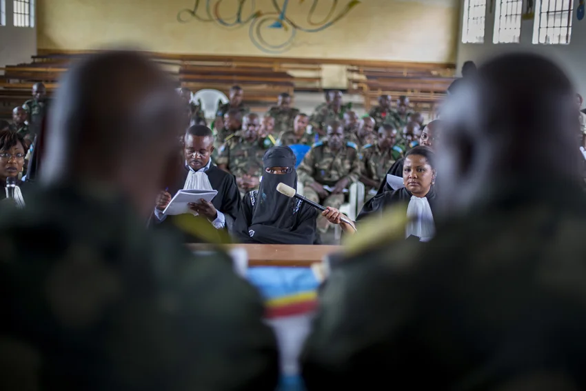 A victim -- veiled to protect her identity -- testifies before the closed military tribunal. A member of the prosecution team holds a microphone up for her while a defense lawyer makes notes. The accused soldiers are seated in the rear. © Diana Zeyneb Alhindawi