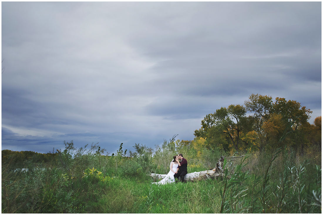 bride and groom kissing on log on overcast day in fall A stunning Fall wedding in Lethbridge Alberta - a religious church ceremony at the URC Church with stained glass windows, a quick stop at the Wooden Shoe for lunch and bridal party pictures at Indian battle park. #albertawedding #lethbridge #redwinewedding #redcolourscheme #babiesbreathboquet #indianbattlepark #woodshoe #overcastwedding #fallwedding #kissingcouple #brideandgroom #emotionalwedding #church #churchceremony
