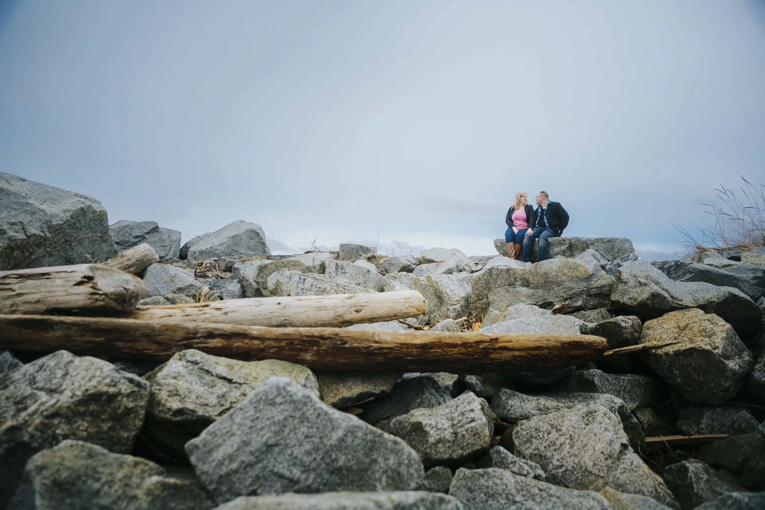 Cloudy engagement session at Garry Point Park in Richmond, BC. Couple sitting on lots of rocks Photography by Christina Voorhorst of Mimsical Photography #westcoastphotographer #engagement #ring #couple #ocean #cloud #overcast #romantic #poses