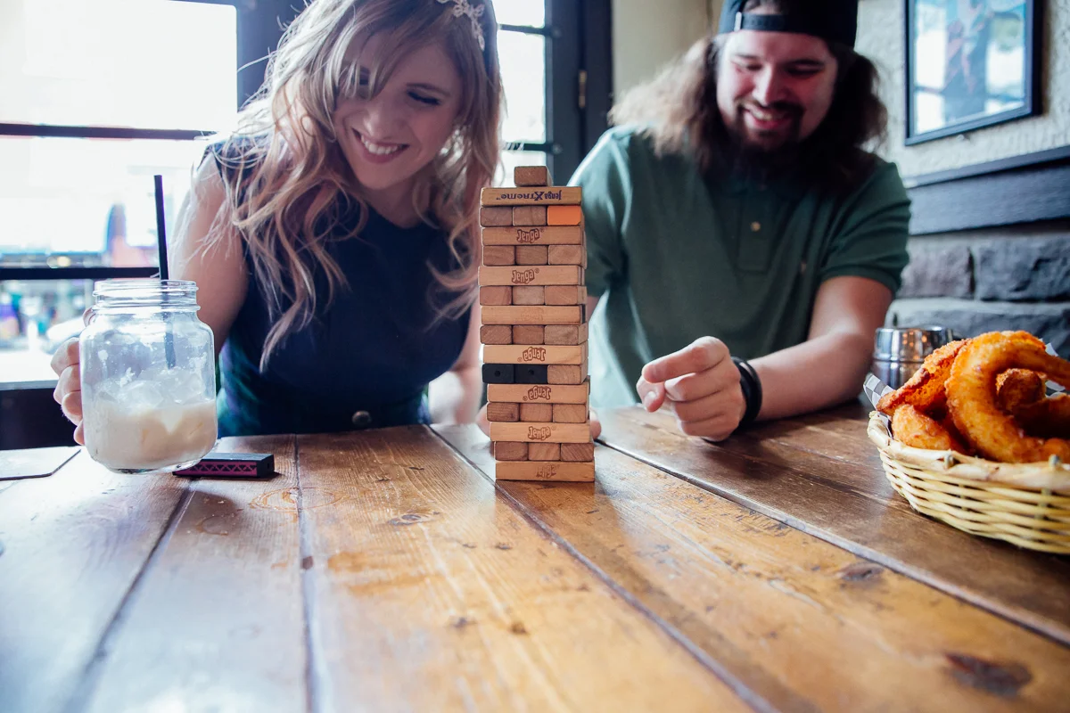 Couple playing Jenga in tavern, butterbeer drink. Genuine Smiles Storm Crow Tavern Documentary Engagement Photography, Nerd Bar, Board Games by Mimsical Photography. Real stories about Real Couples that are just as unique as they are. #unique #vibrant #personality #nerd #princess #ecclectic #gamer #couple #tavern #vancouver #candid #storytelling