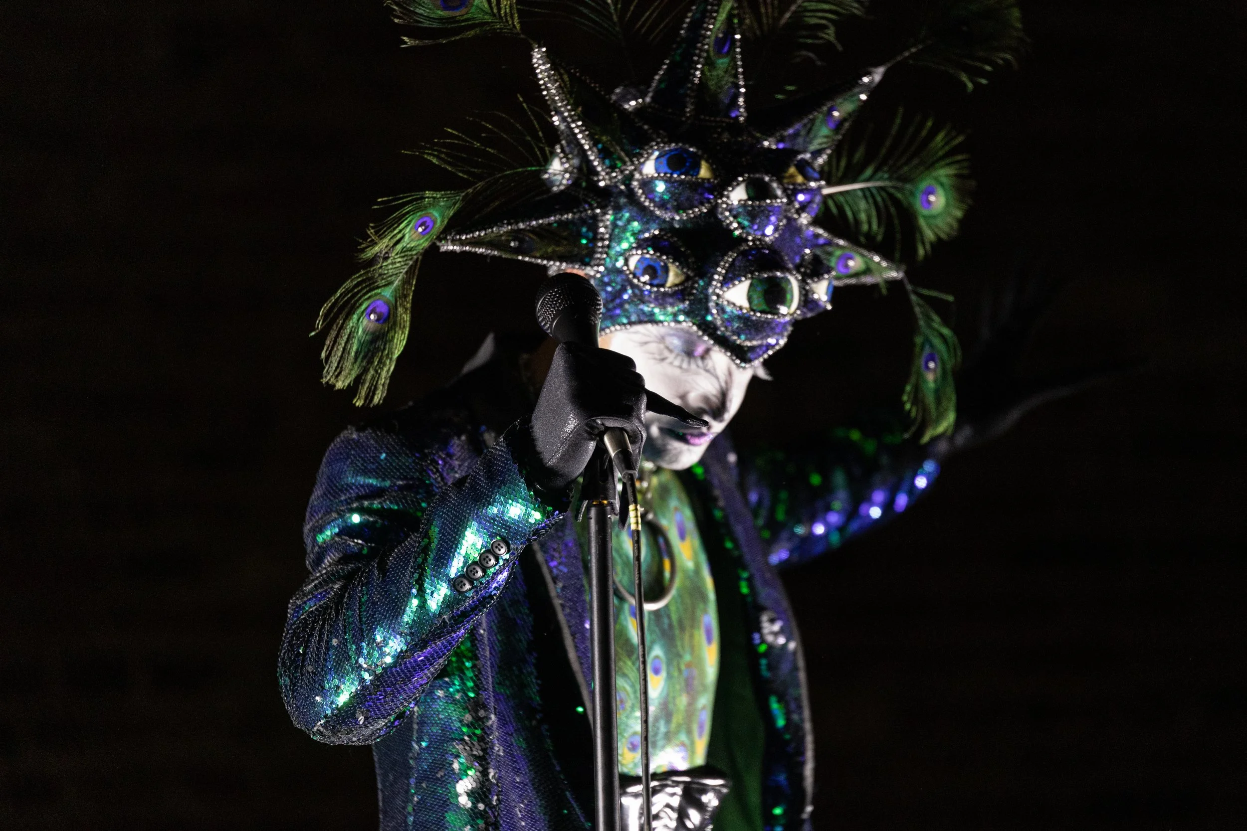 Performer in elaborate peacock-themed costume with peacock feather headpiece, black gloves, and sequined blazer, holding a microphone against a dark background.