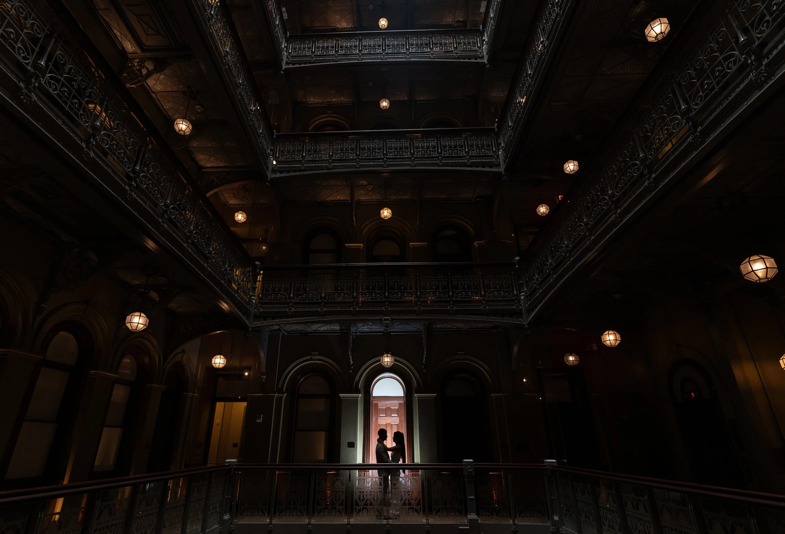 Silhouettes of a couple facing each other inside a grand, dark, multi-story building with ornate railings and hanging lights, backlit by a bright doorway.