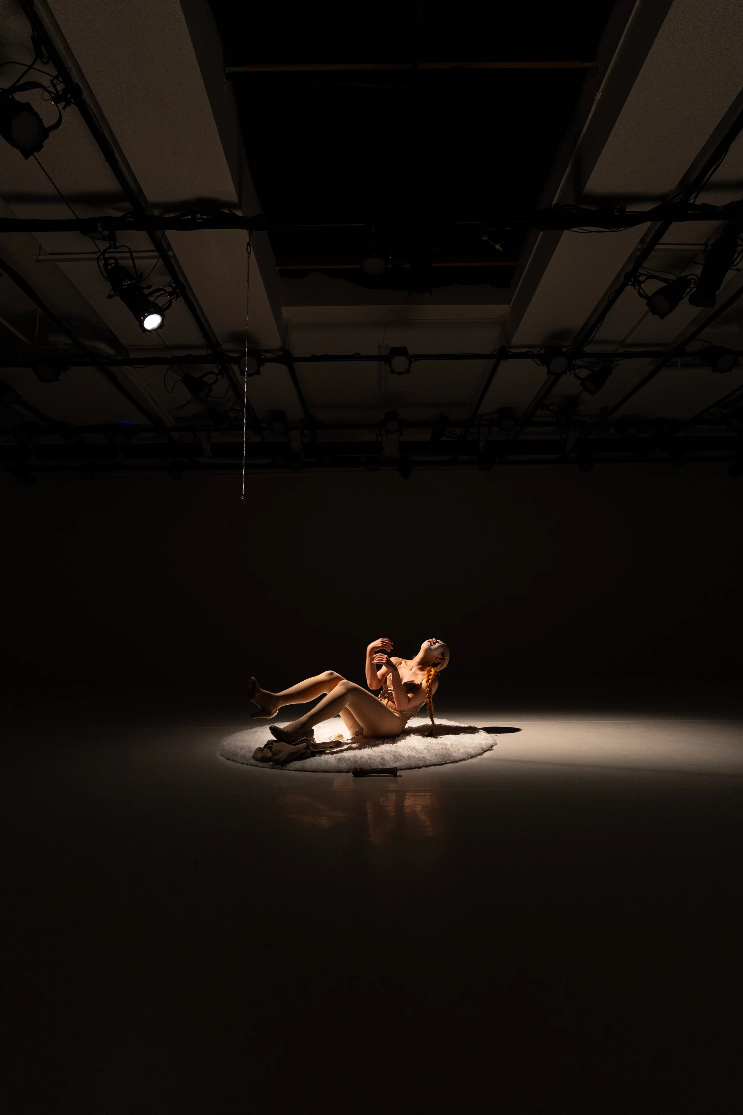 A person lying nude on a round rug in a spotlighted area, with their head tilted back and arms bent.