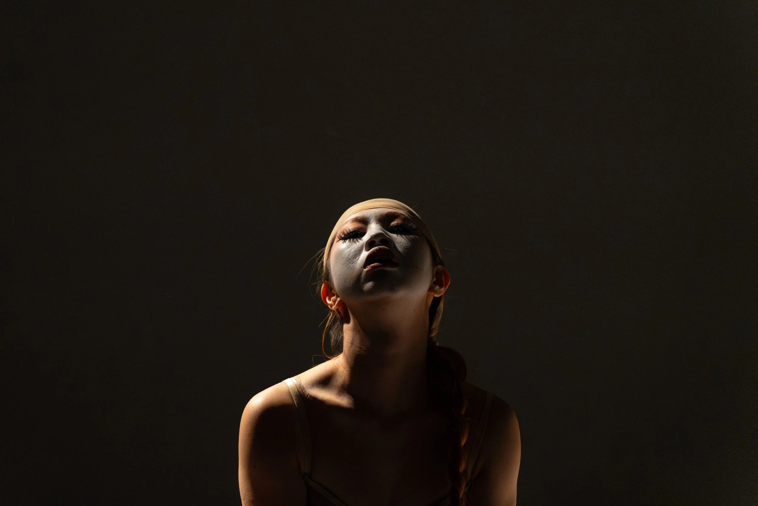 A woman with makeup and long hair, wearing a headband, looking upward with a serious expression, against a dark background.
