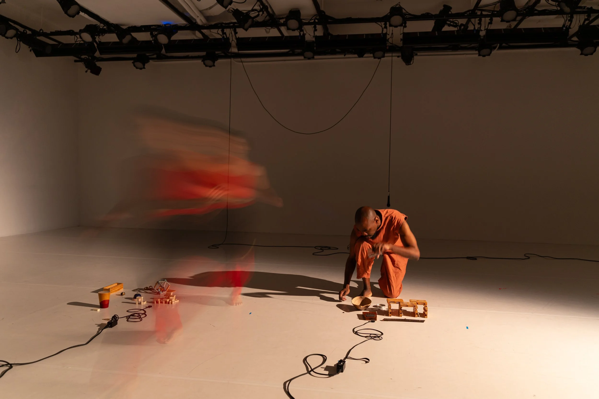A person dressed in orange crouching on the floor of a stage or studio, surrounded by various objects and wires, with a blurred figure moving rapidly nearby, and a shadow cast on the wall behind.
