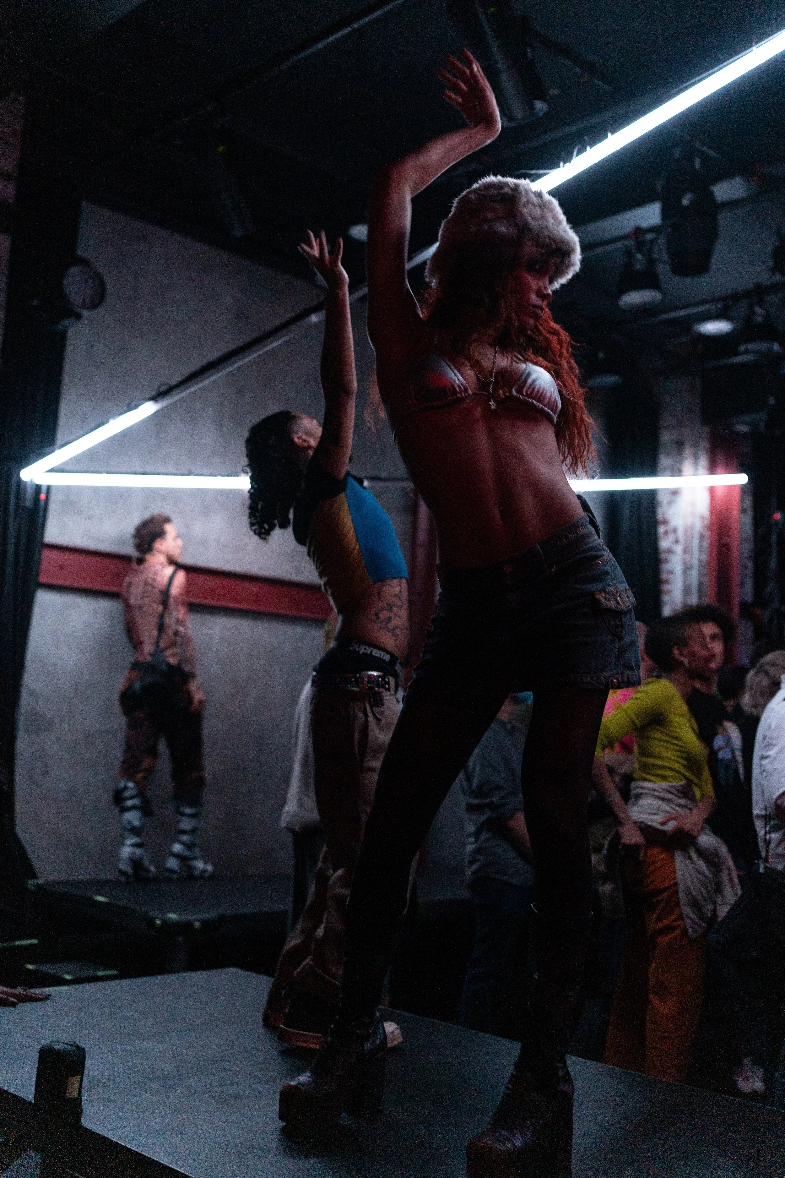 People dancing on a stage in a dark club with neon lighting, including a woman in crop top and platform shoes in the foreground.