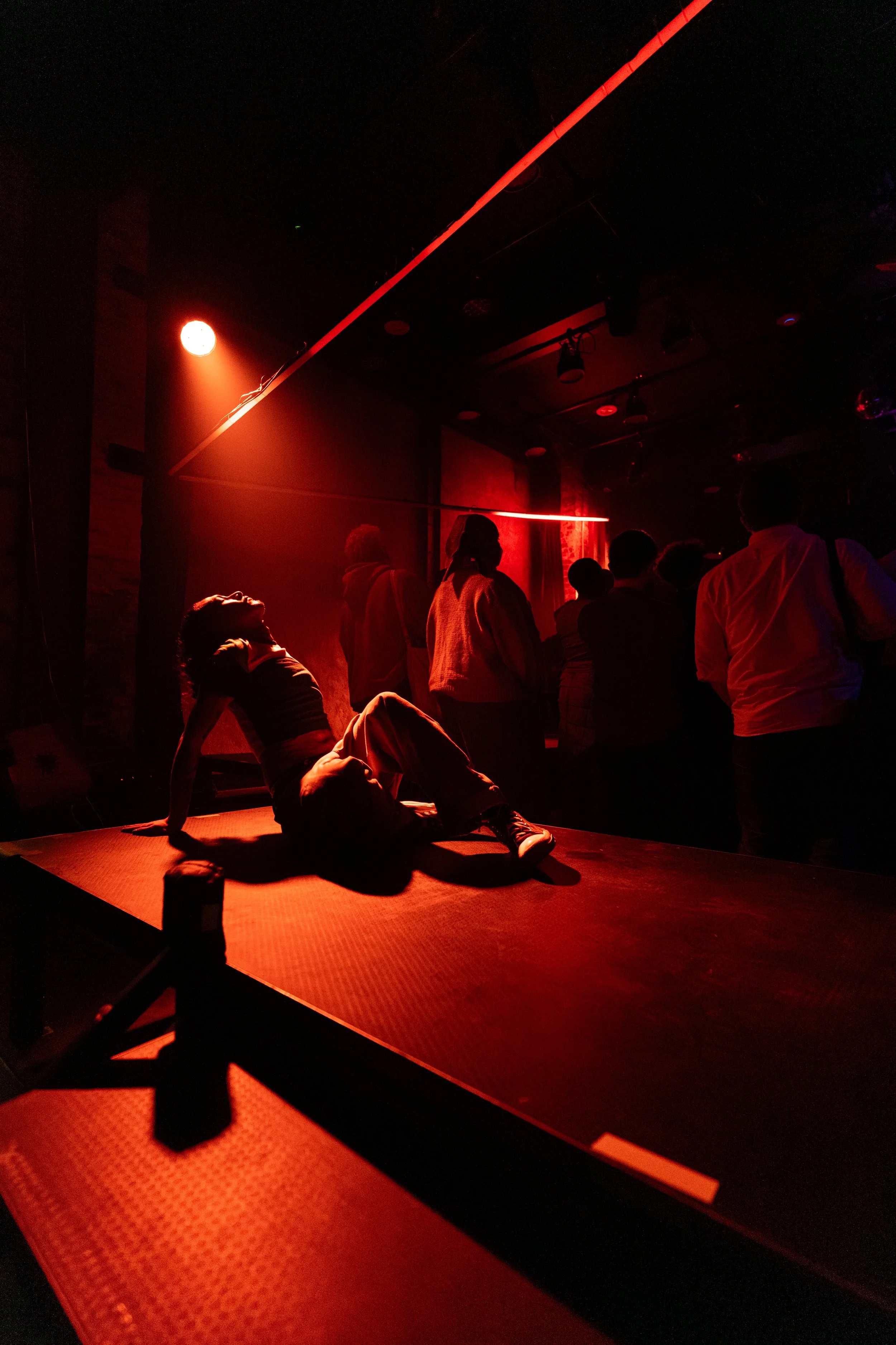 People in a dimly lit entertainment venue with red lighting, including a woman sitting on a table, and others standing and socializing in the background.