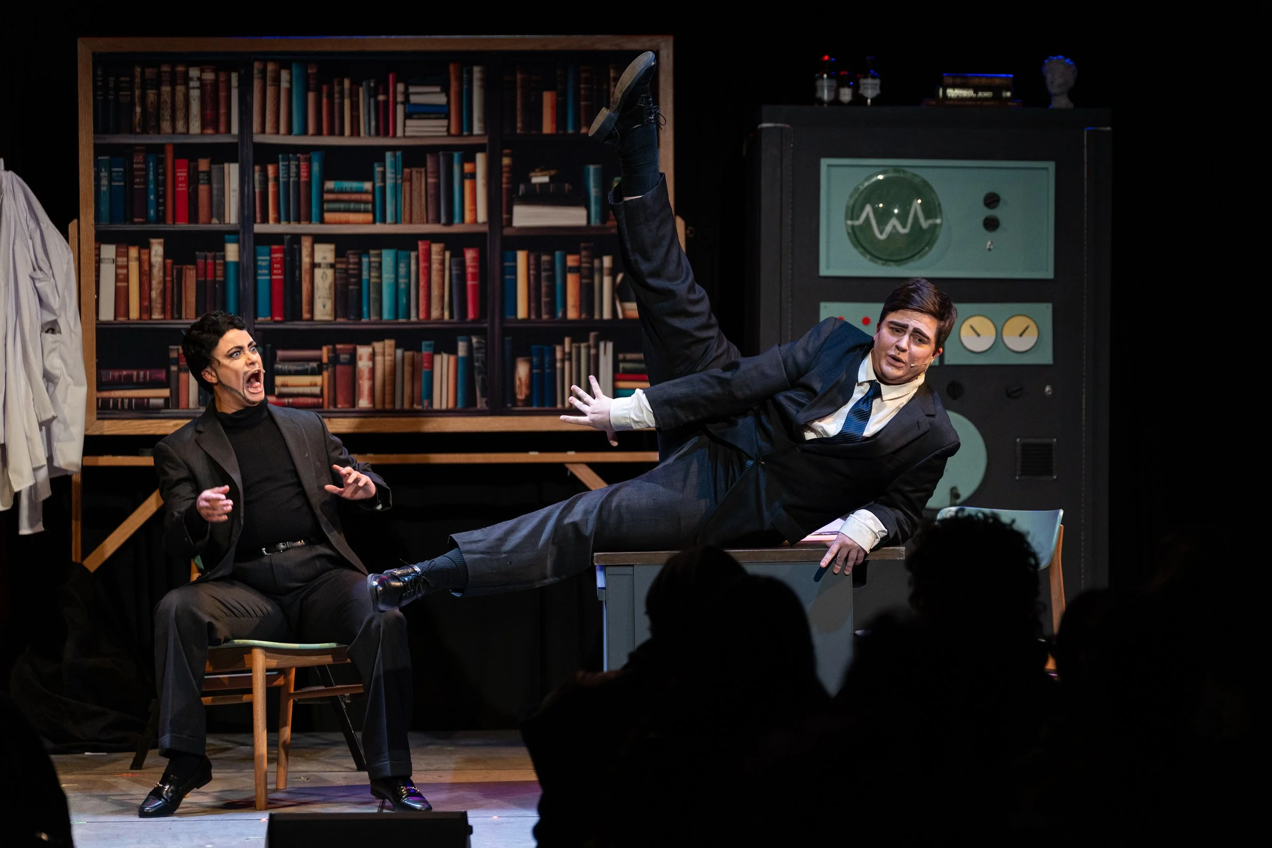 Stage scene with two actors in a theatrical performance, one woman seated and yelling, another man in a suit flying through the air with a surprised expression, set with bookshelves and vintage scientific equipment.
