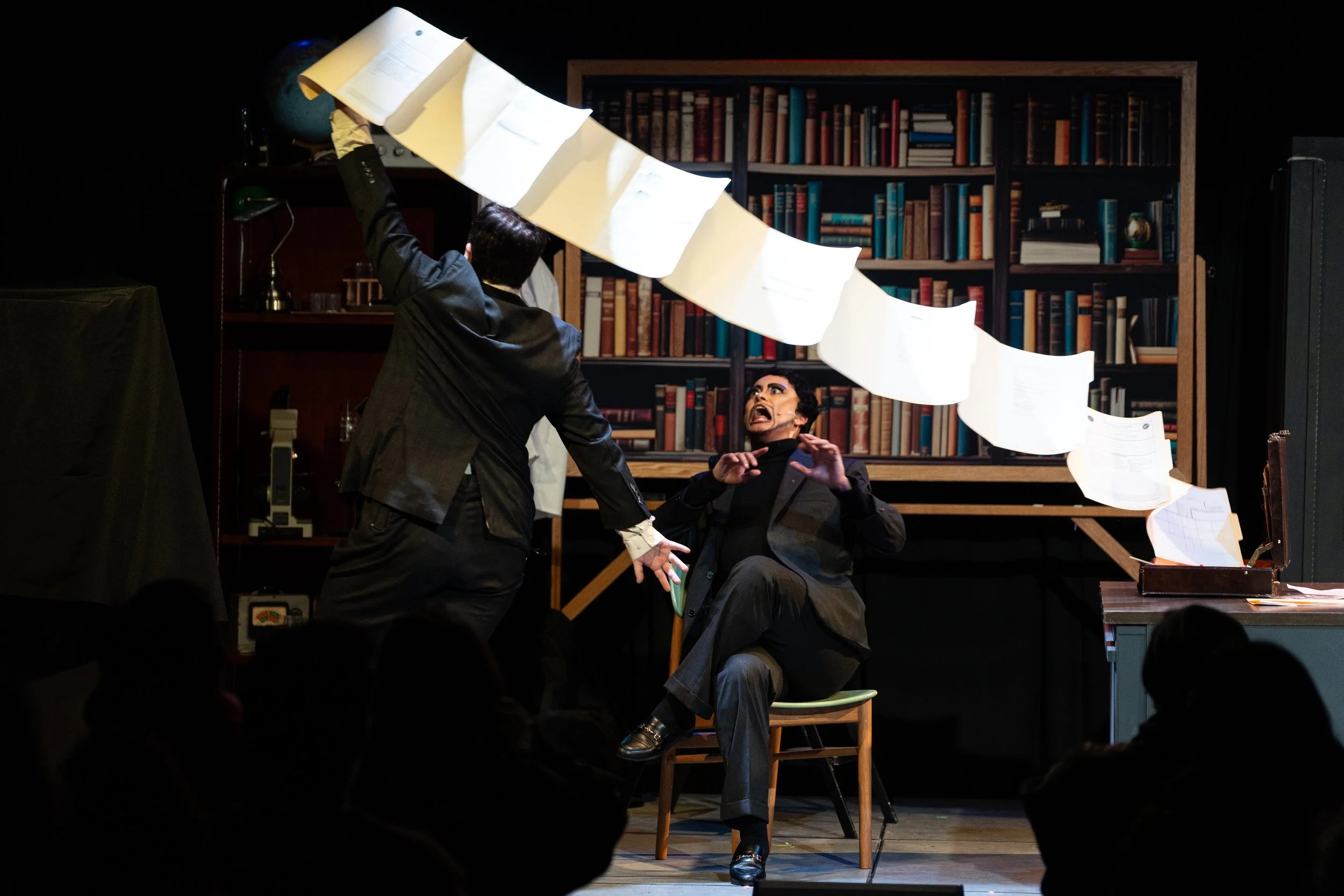 The image depicts a theatrical performance featuring two actors on stage. One actor is standing, raising their arm with papers or scripts hanging in the air, while the other actor is seated, making an animated expression with hands raised. The backgr