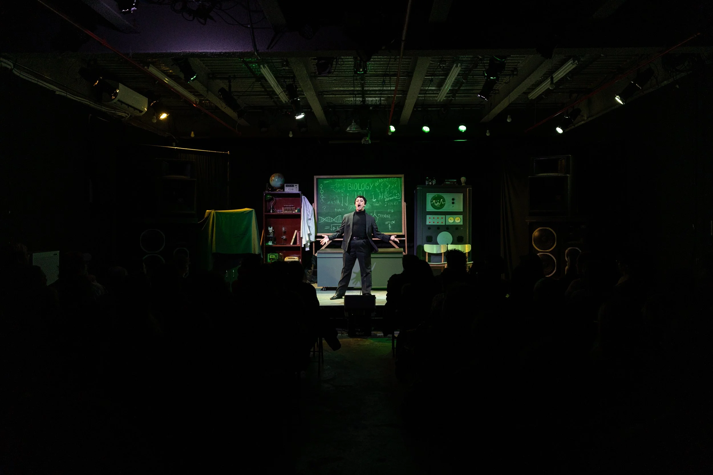 A person is performing a monologue or presentation on a stage designed to look like a science classroom, with a chalkboard, bookshelves, and science equipment.