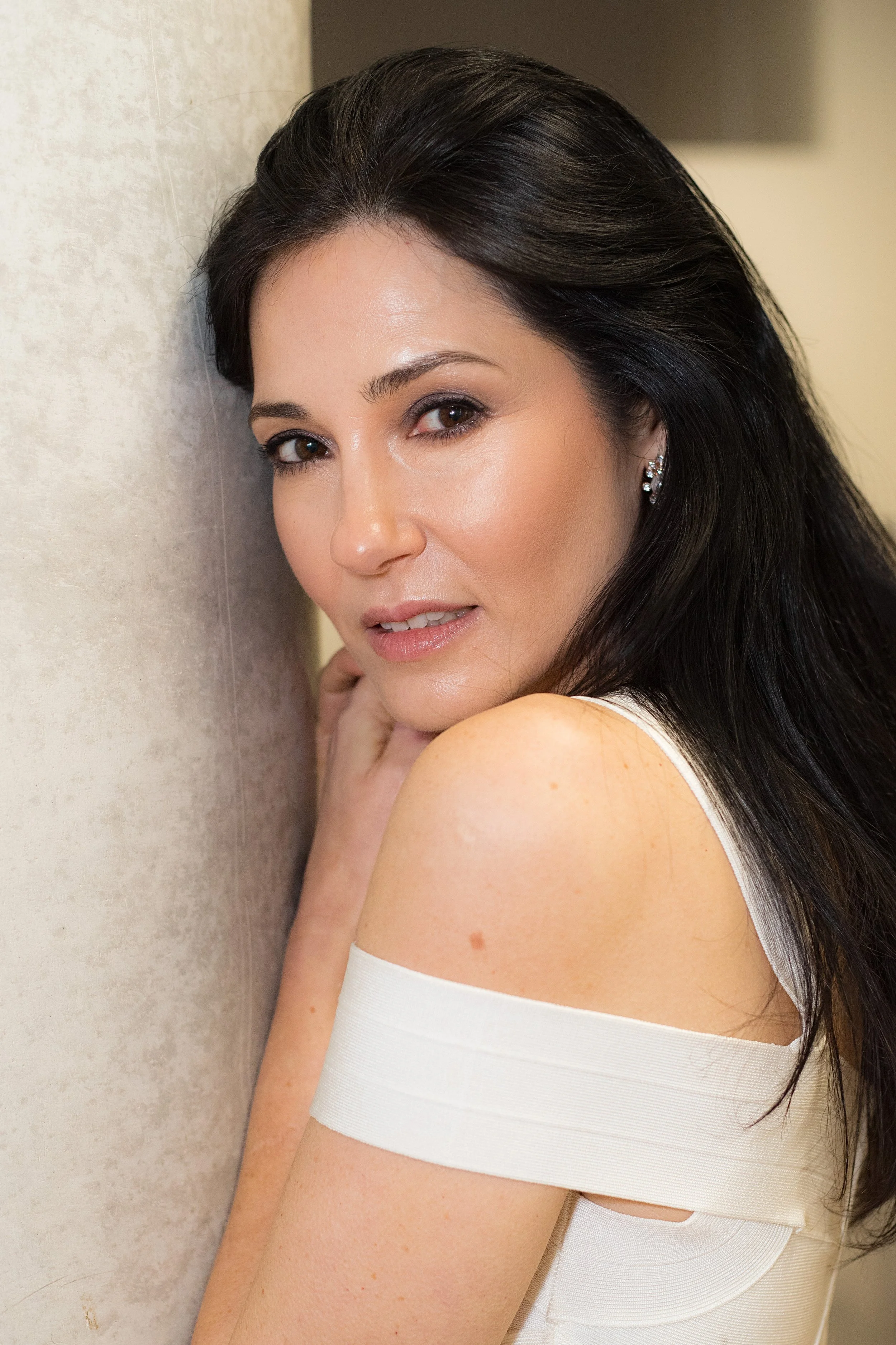 Close-up of a woman with long dark hair, wearing earrings and a white off-shoulder top, leaning against a wall and looking at the camera.